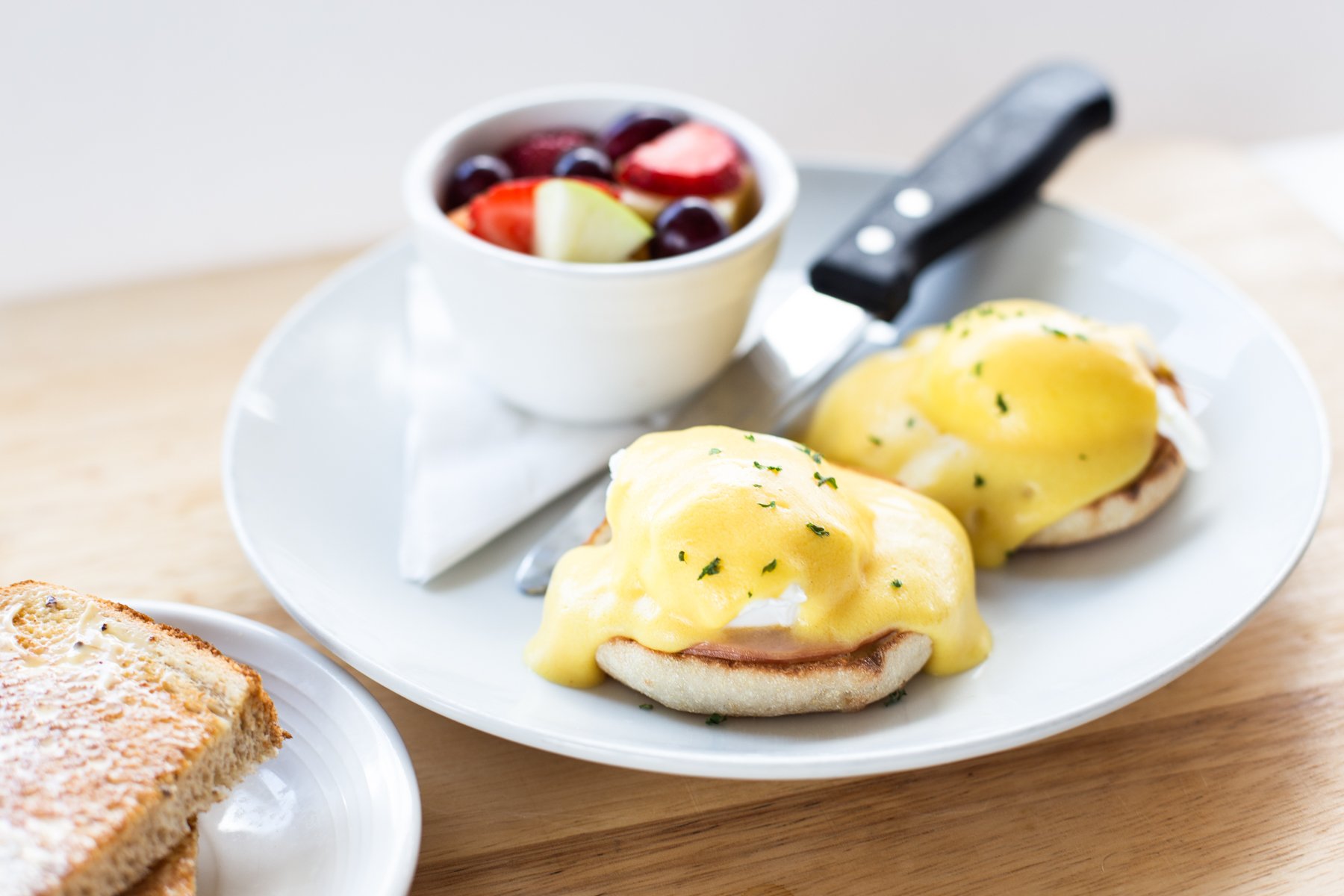 Eggs Benedict with hollandaise sauce and chopped herbs, served on toasted English muffins, with a side of fruit salad containing strawberries, grapes, and other fruits.