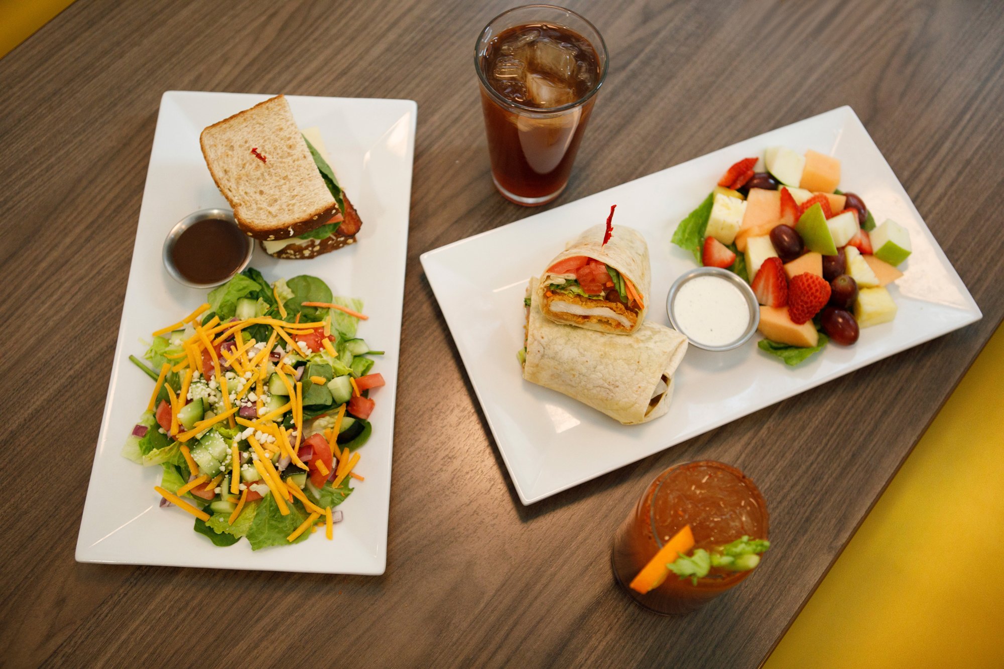 A meal with a salad, a sandwich, a wrap, and a fruit salad on white plates, with two glasses of iced tea on a wooden table.