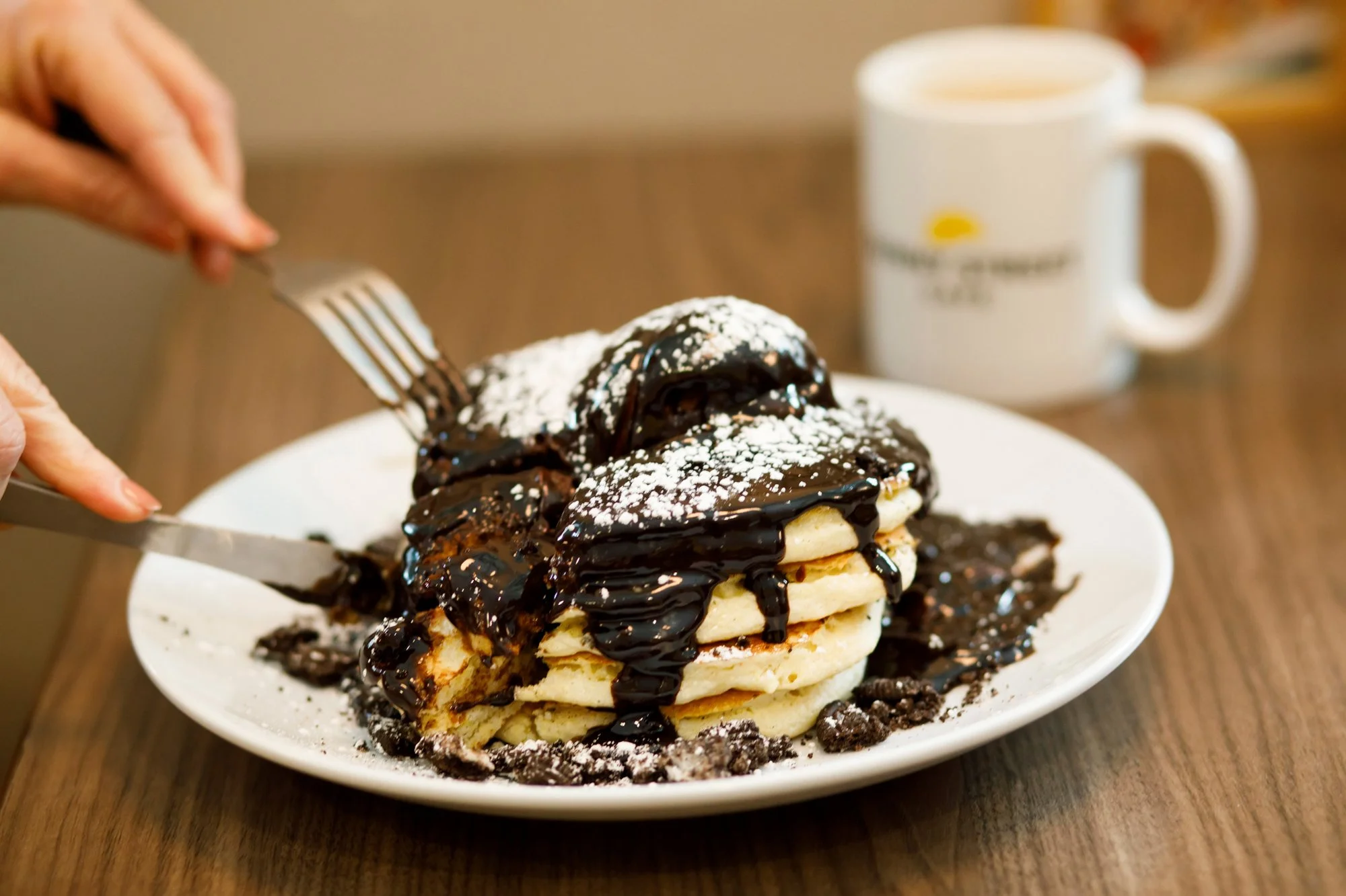 Stacked pancakes topped with chocolate sauce, whipped cream, powdered sugar, and sprinkles, served on a white plate with a fork and a mug of coffee in the background.