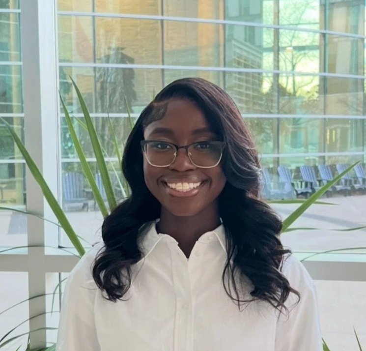 A smiling woman with glasses and long dark hair, wearing a white shirt, standing indoors near large windows and green plants.