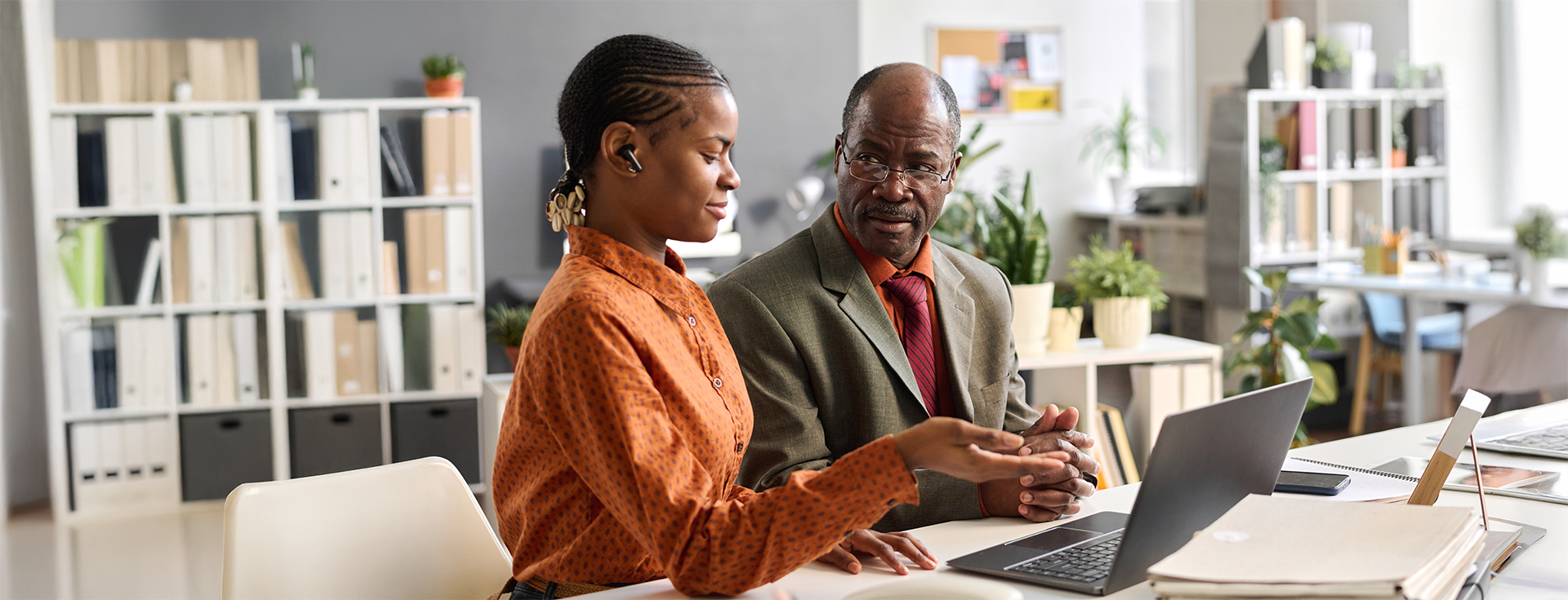 Two professionals, a woman and a man, having a discussion in an office. The woman is speaking with her hand raised and the man is listening with a serious expression. The office background features shelves with books and plants.
