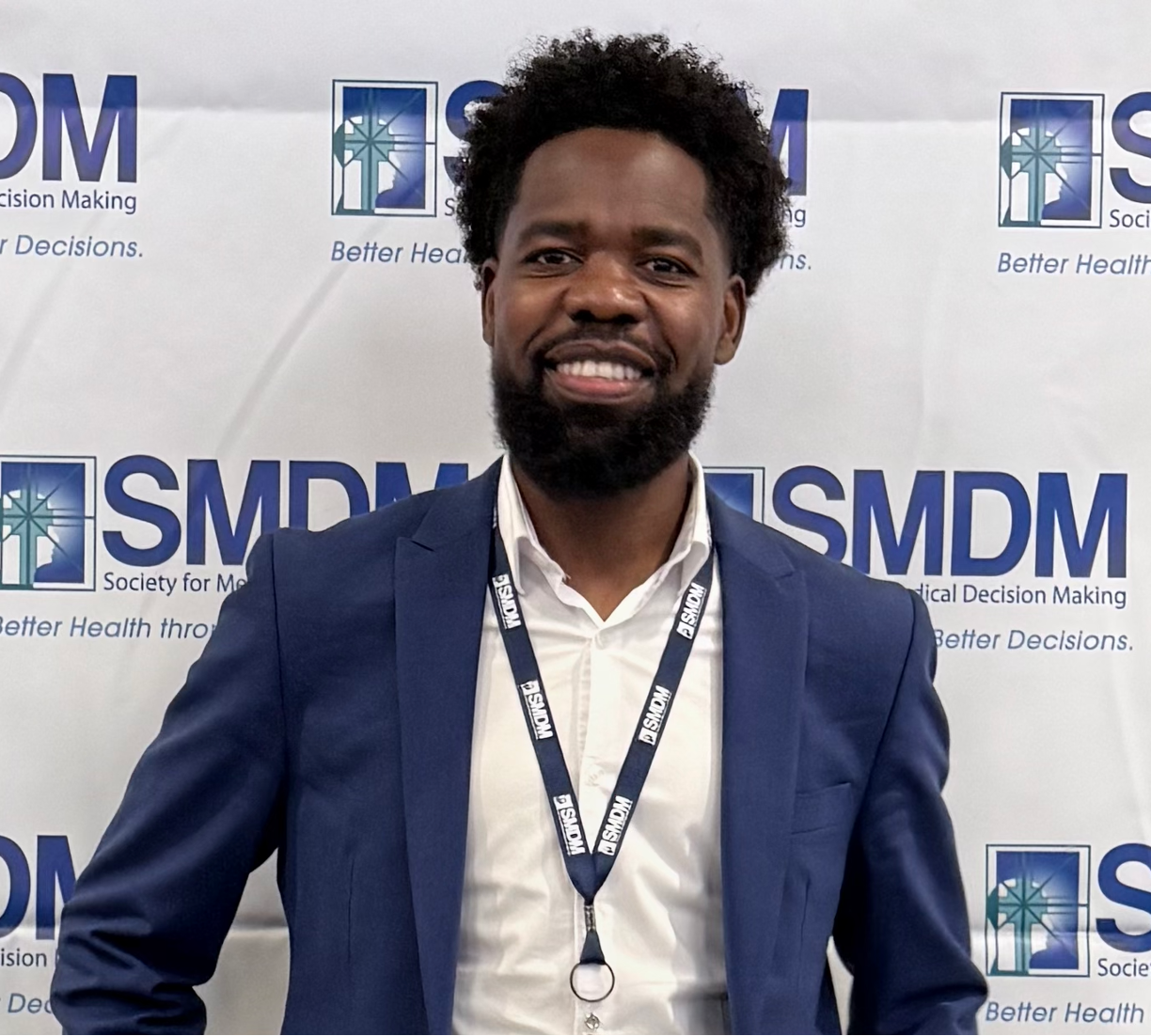 A smiling man with a beard wearing a navy blazer and white shirt, standing in front of a backdrop with logos for the Society for Medical Decision Making (SMDM) and the slogan "Better Health through Better Decisions."