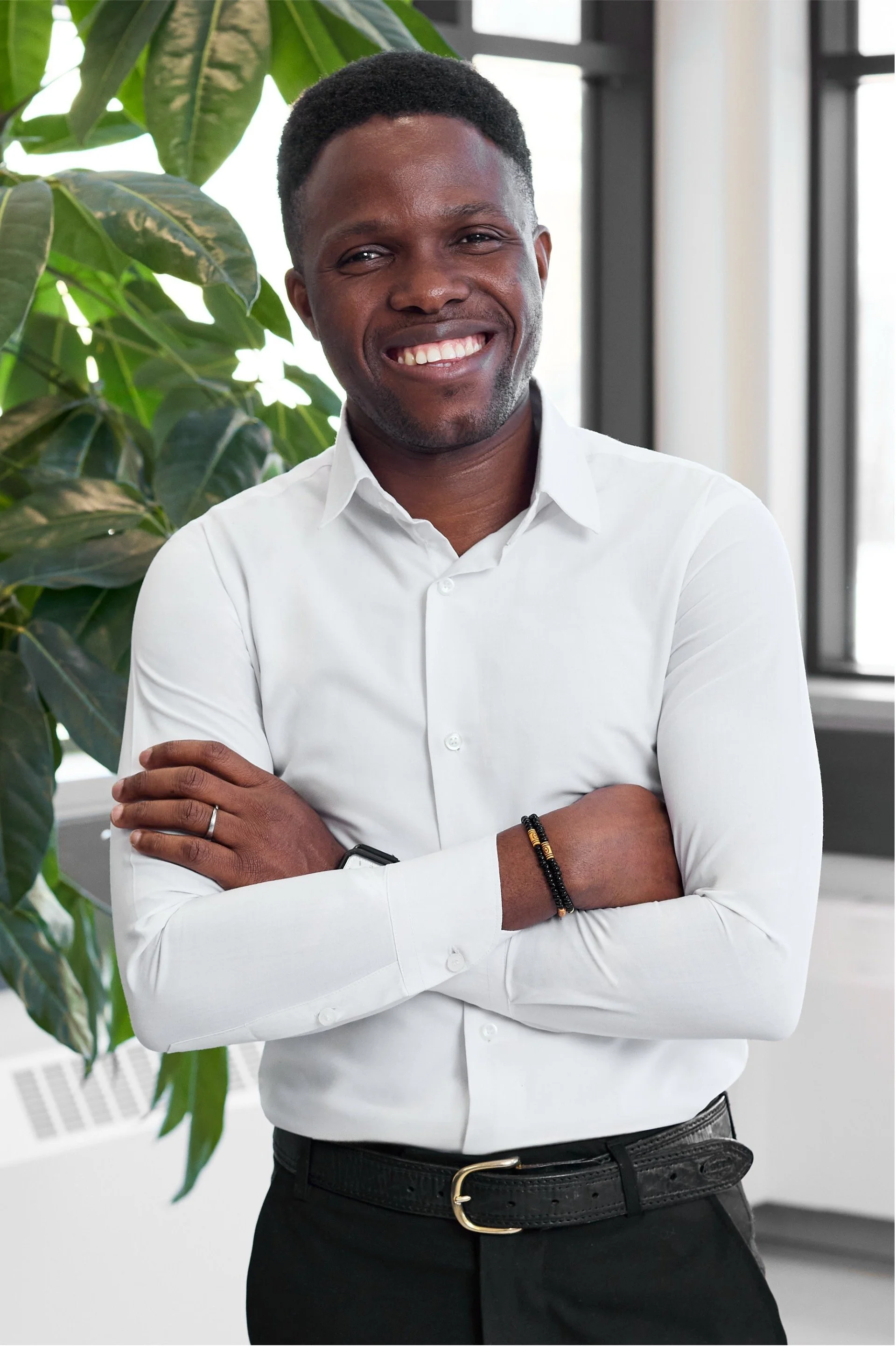 A smiling man with dark skin and short curly hair, wearing a white dress shirt and black pants, standing with arms crossed in a modern indoor setting with large windows and green plants.