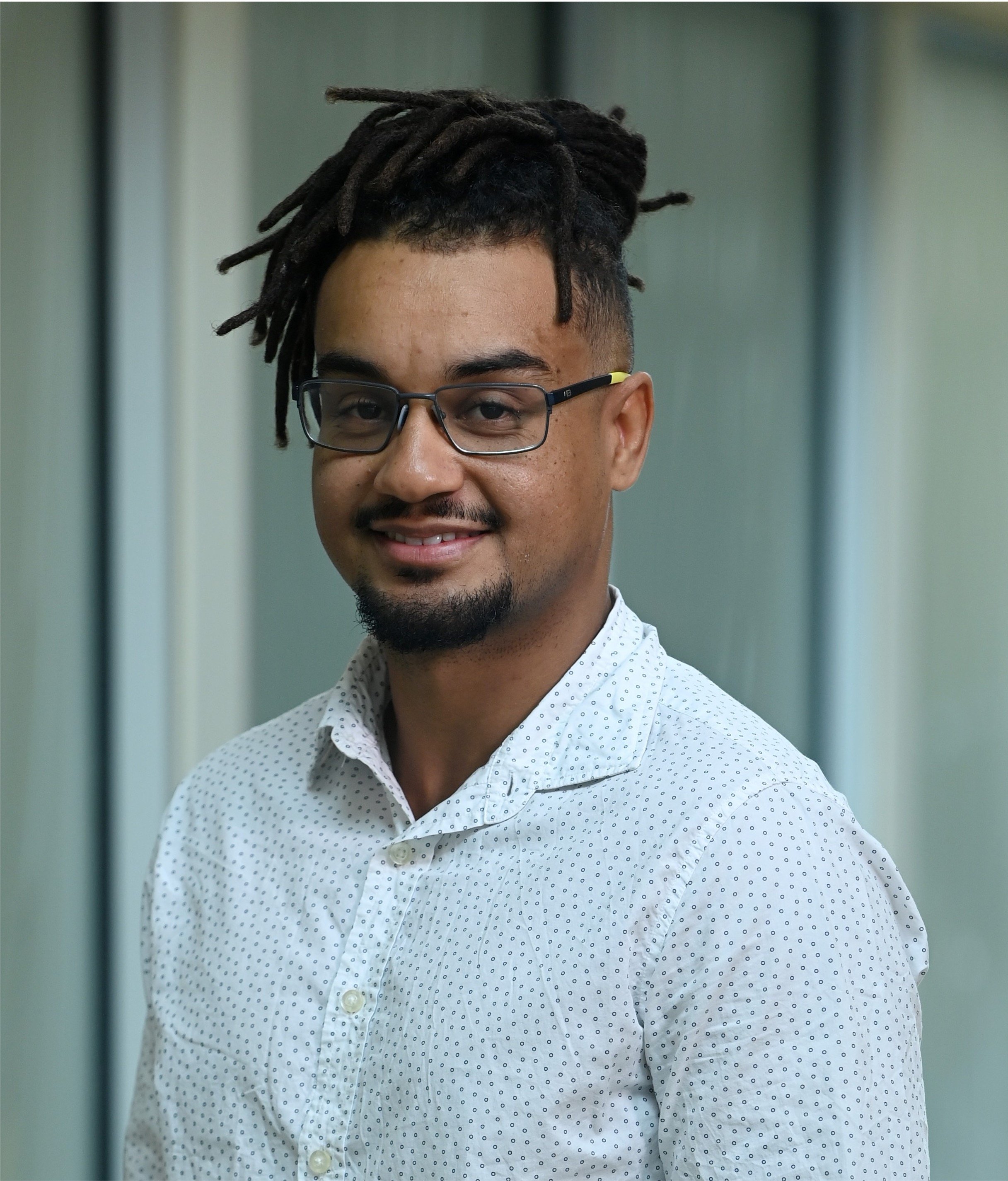 A smiling man with glasses, dark dreadlocks, and a goatee, wearing a white shirt with small black dots, standing indoors.