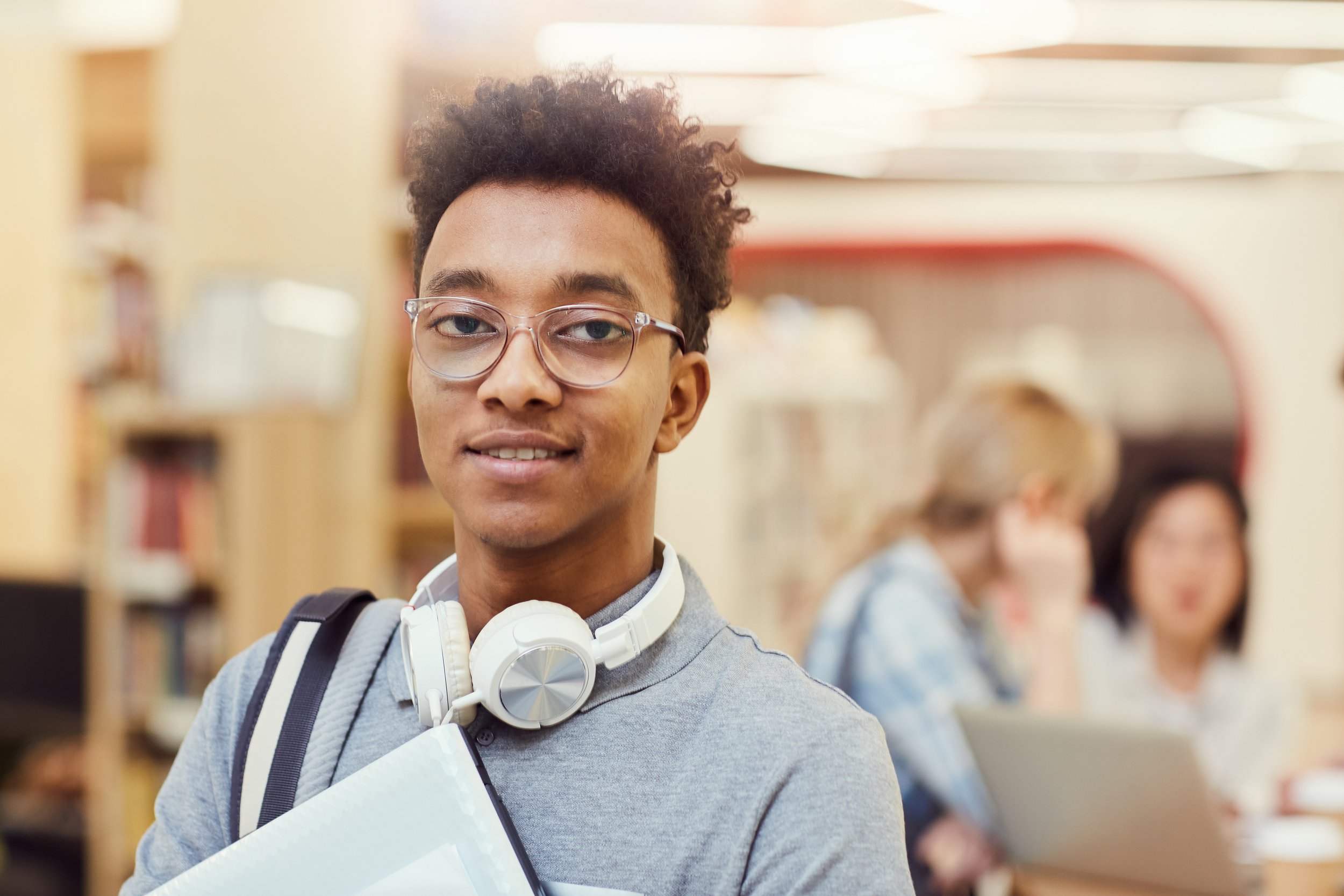 Young man with curly hair, glasses, and headphones around his neck standing in a library, with bookshelves and blurred people in the background.