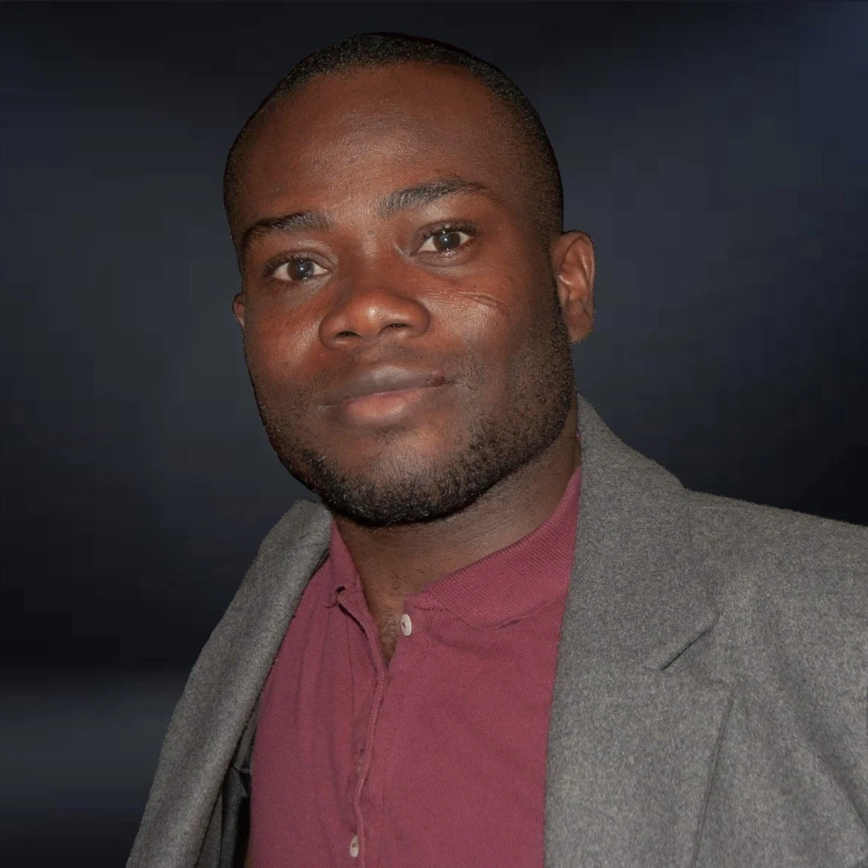 A professional headshot of a Black man with short hair and a beard, wearing a maroon shirt and a gray blazer, standing against a dark background.