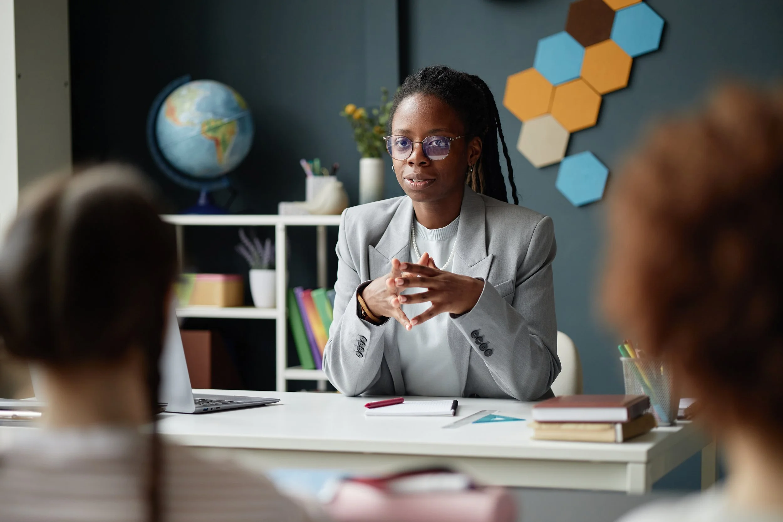An African American woman with glasses, wearing a gray blazer, sitting at a desk in a classroom or office setting, engaging in conversation with children.