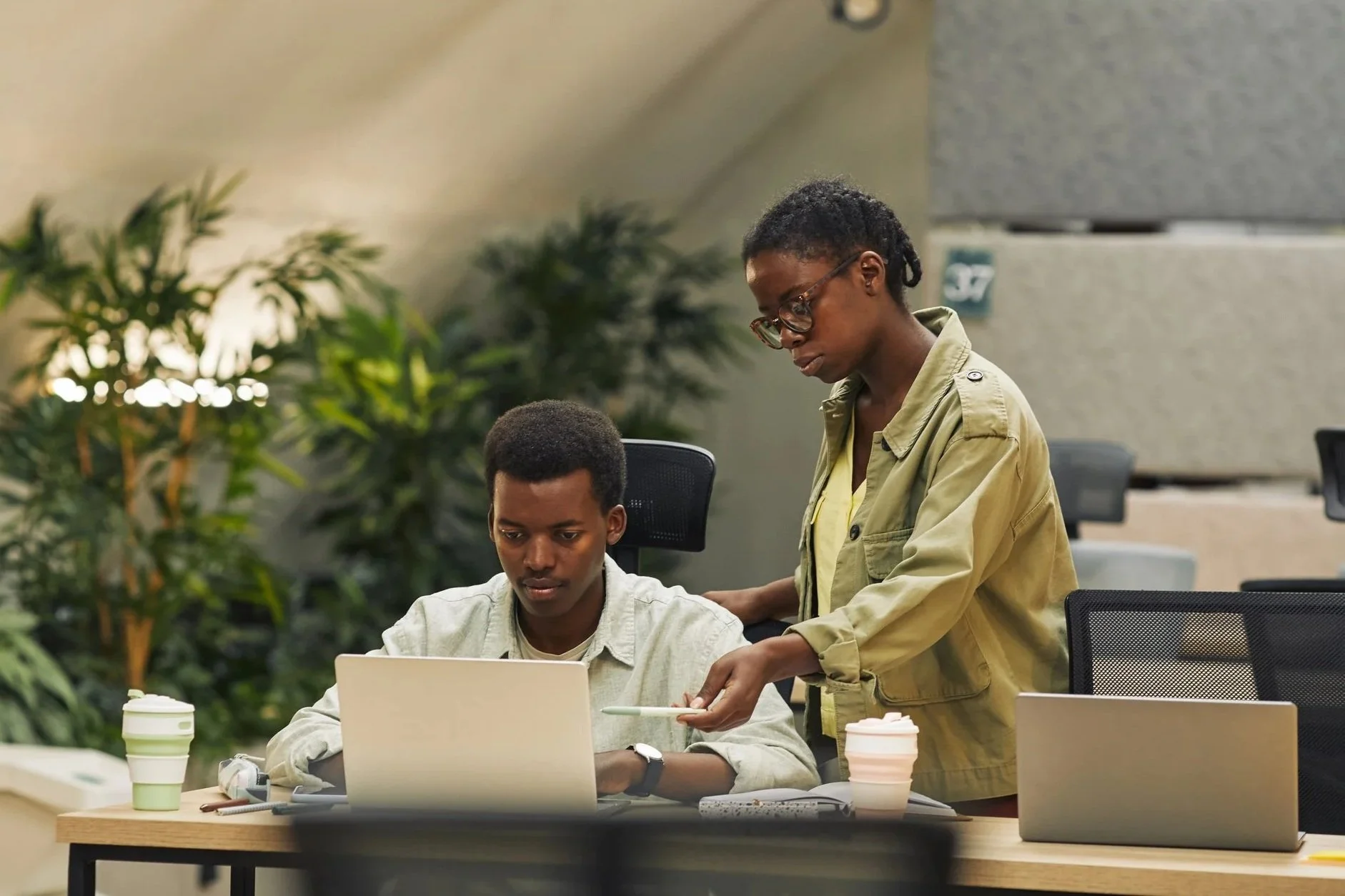 Two young men are working together in an office filled with plants. One man is sitting at a table with a laptop, and the other man is standing beside him, handing him a tablet. There are coffee cups and notebooks on the table.