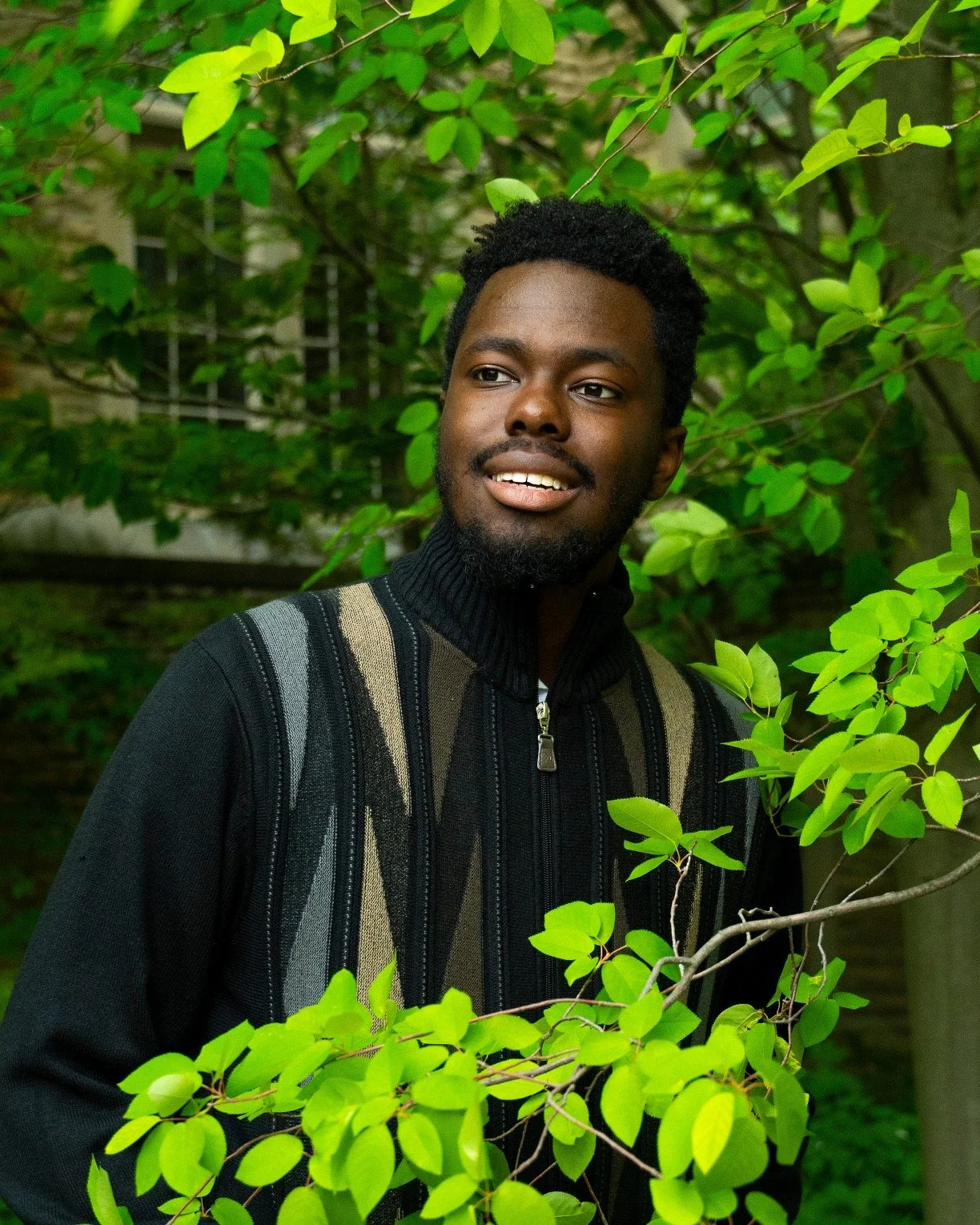 Portrait of a young man with dark hair and a beard, smiling slightly, surrounded by green leaves and trees.