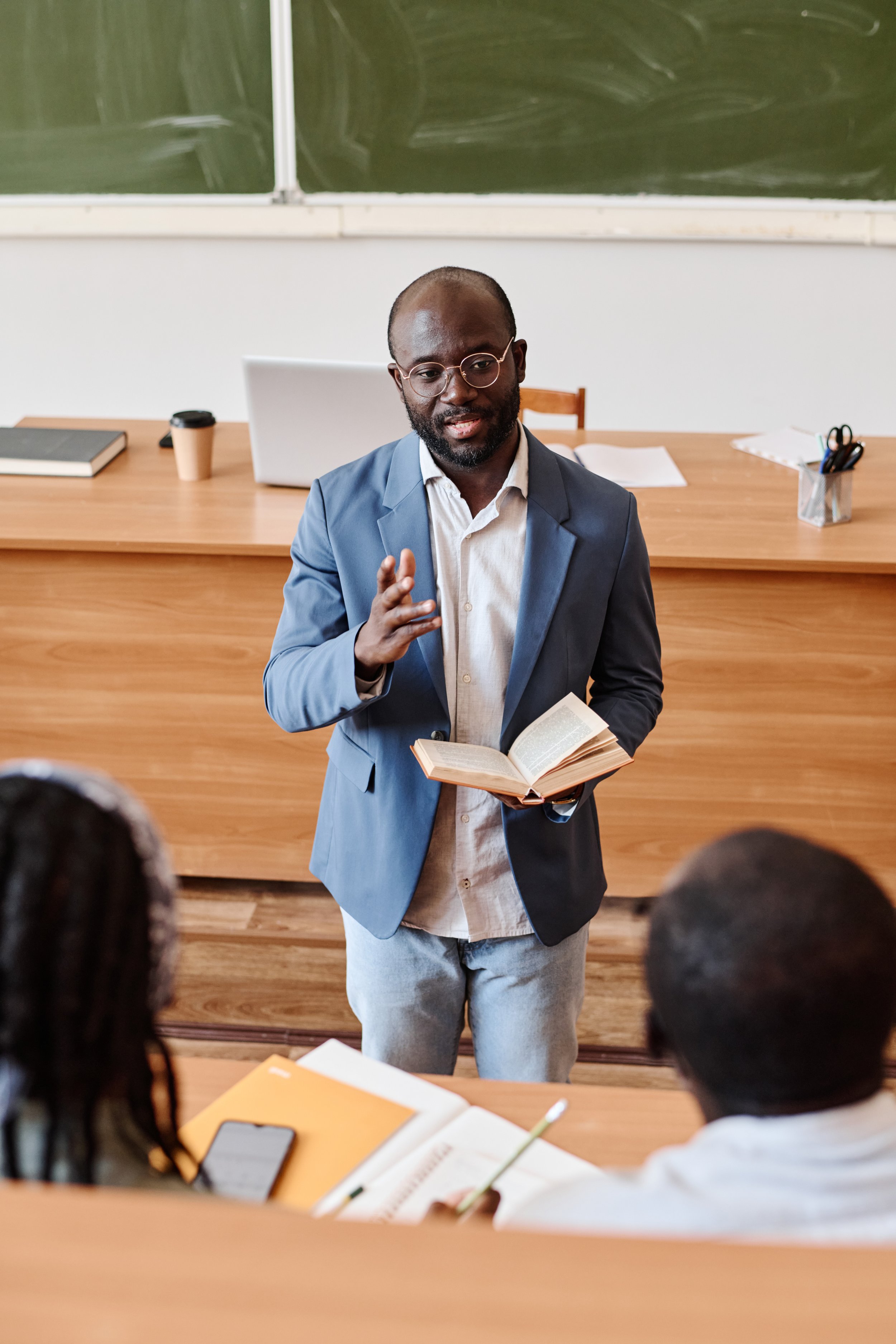 A man in a blue blazer and glasses is giving a lecture or presentation in a classroom, holding a book, with students seated in front of him and a blackboard behind.