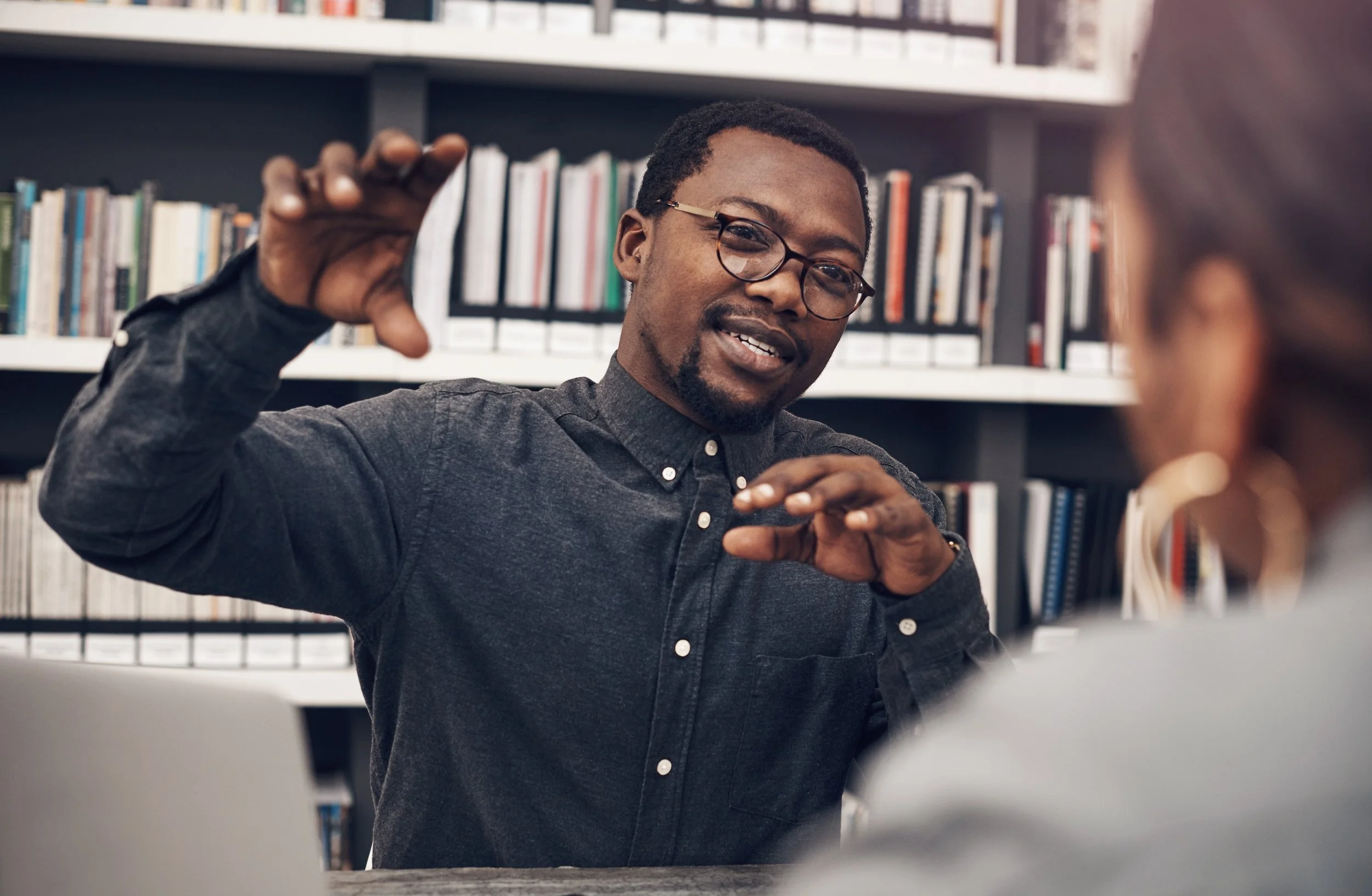 A young man with glasses and a beard, wearing a dark button-up shirt, is smiling and gesturing with his hands in a lively conversation in a library with bookshelves in the background.
