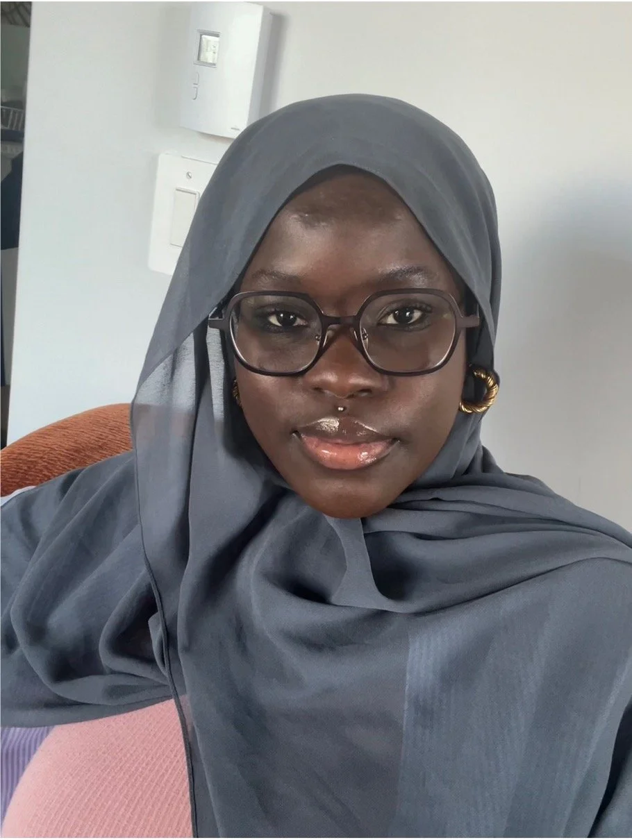 A woman wearing a gray headscarf and large eyeglasses, sitting indoors on a pink cushioned chair with a neutral expression.