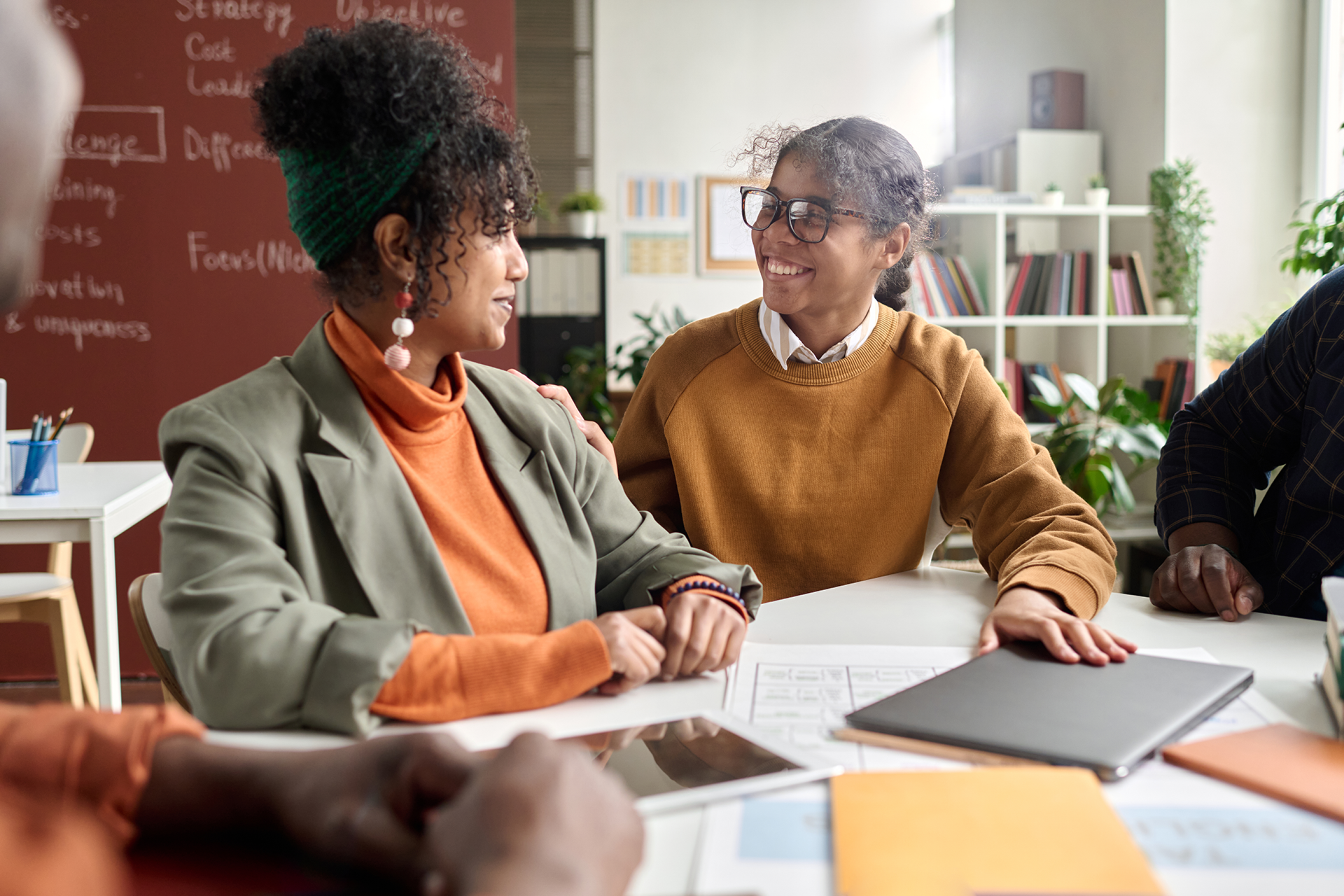 Two women talking and smiling during a meeting or discussion at a table in an office setting, with bookshelves and plants in the background.