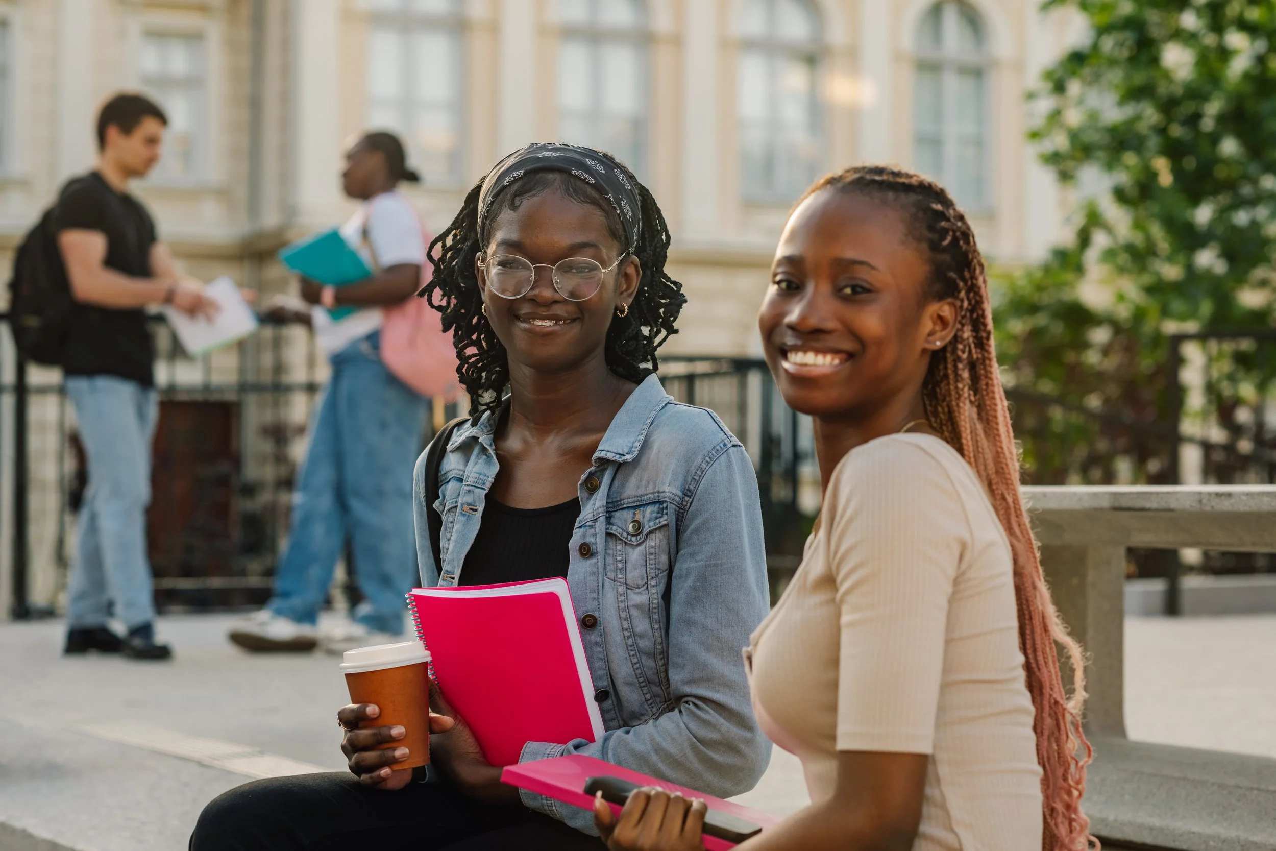 Two smiling young women sitting together outdoors, one holding a pink notebook and coffee, with study partners in the background.