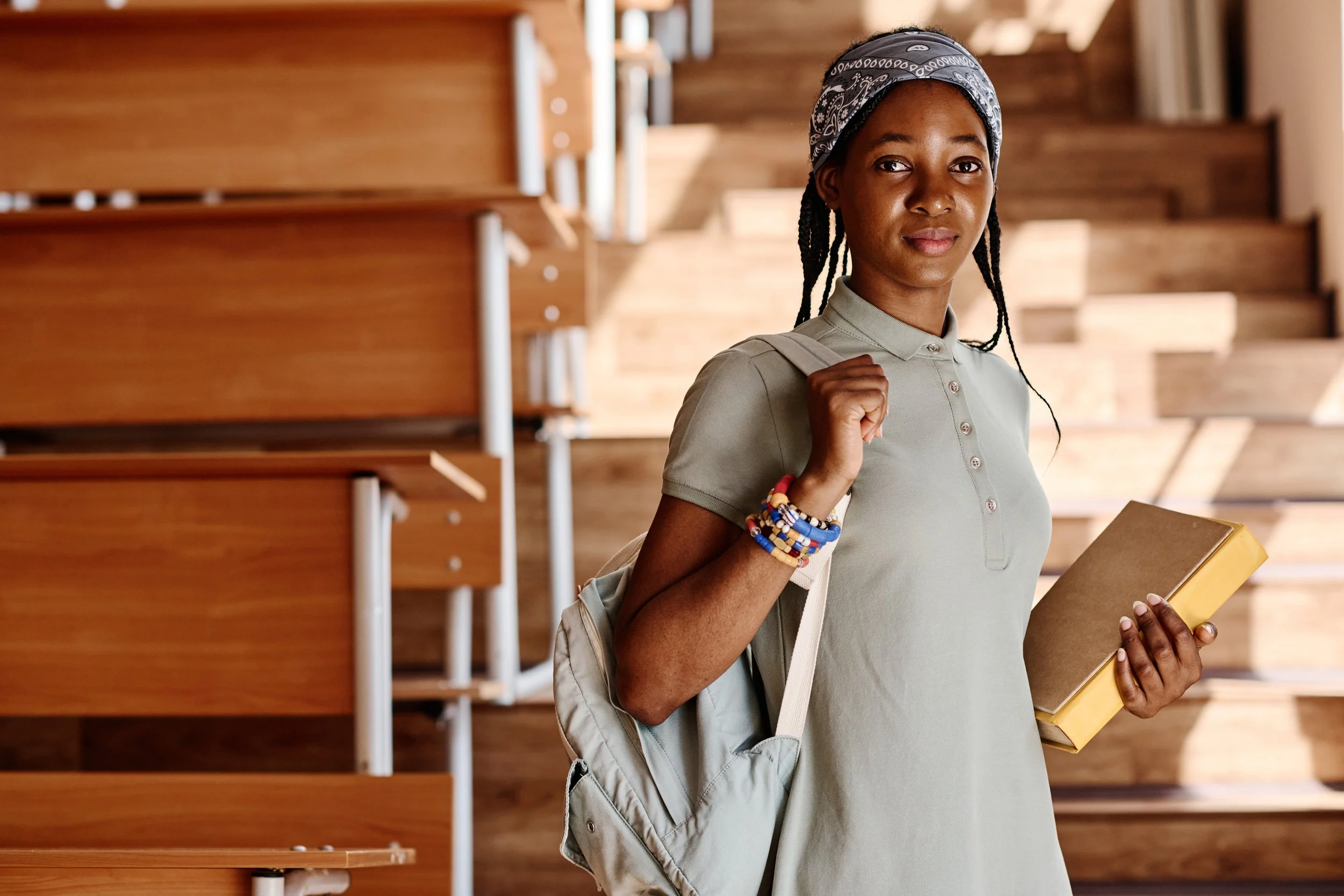 A young African American woman with braided hair, wearing a grey polo shirt, a blue bandana, multiple colorful bracelets, and a light jacket over her shoulder, standing inside a wooden staircase with a warm tone. She is holding a yellow notebook and carrying a grey backpack, looking at the camera.
