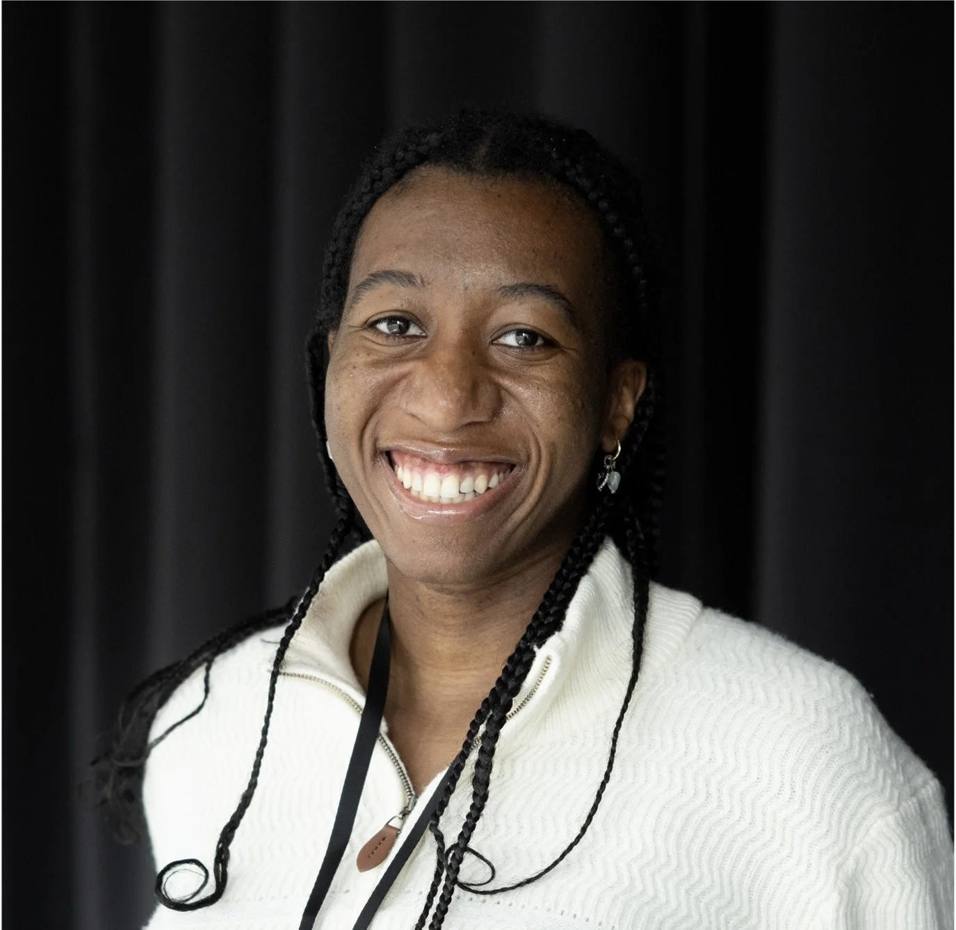 Close-up of a smiling woman with dark braided hair wearing a white sweater and earrings, standing in front of a dark background.