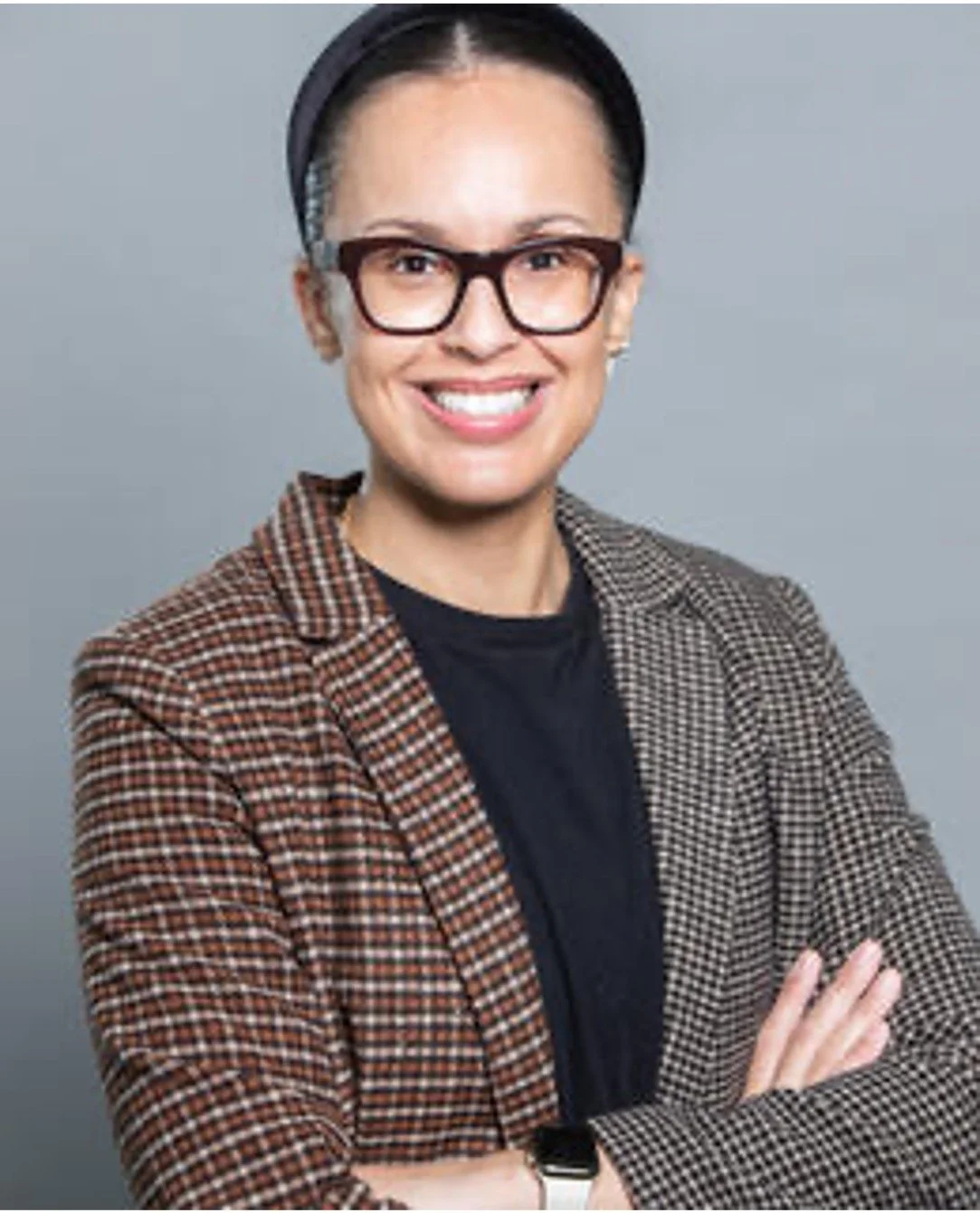 Woman smiling with glasses, wearing a plaid blazer and black shirt, with arms crossed, against a gray background.