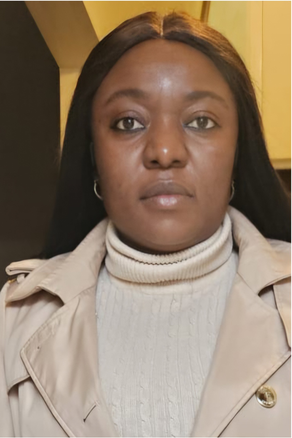 Close-up of a woman with dark hair, wearing a beige trench coat and a cream turtleneck sweater, indoors with warm lighting.