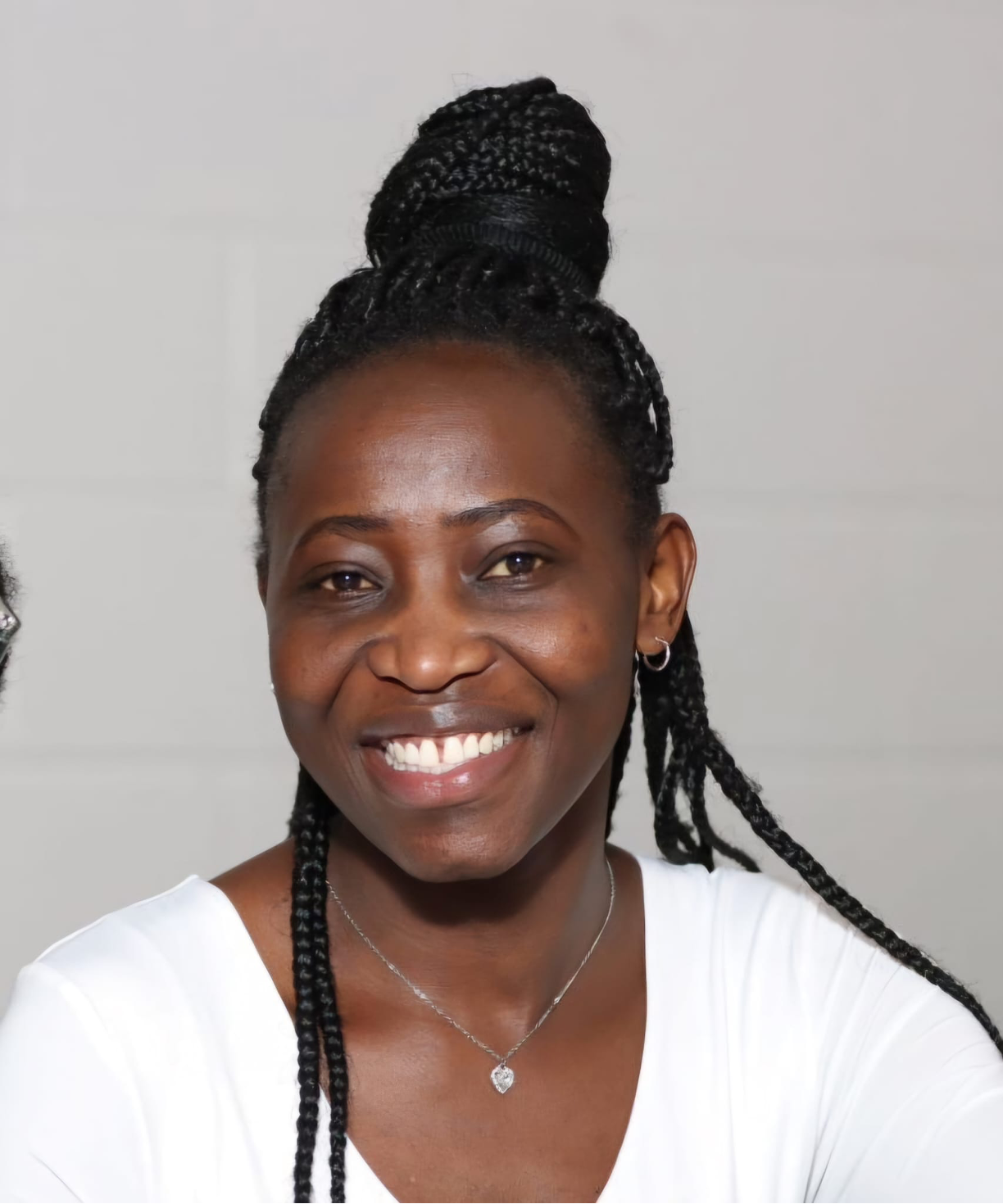 Close-up of smiling woman with braided hair styled in a high bun, wearing hoop earrings, a necklace with a heart pendant, and a white top, against a plain background.