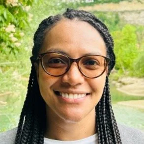 A woman with glasses and braids smiling outdoors with greenery and water in the background.