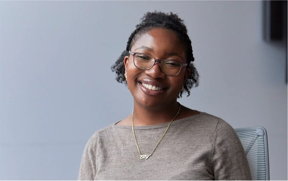 A woman with glasses and curly hair smiling and sitting in a gray chair against a plain background.