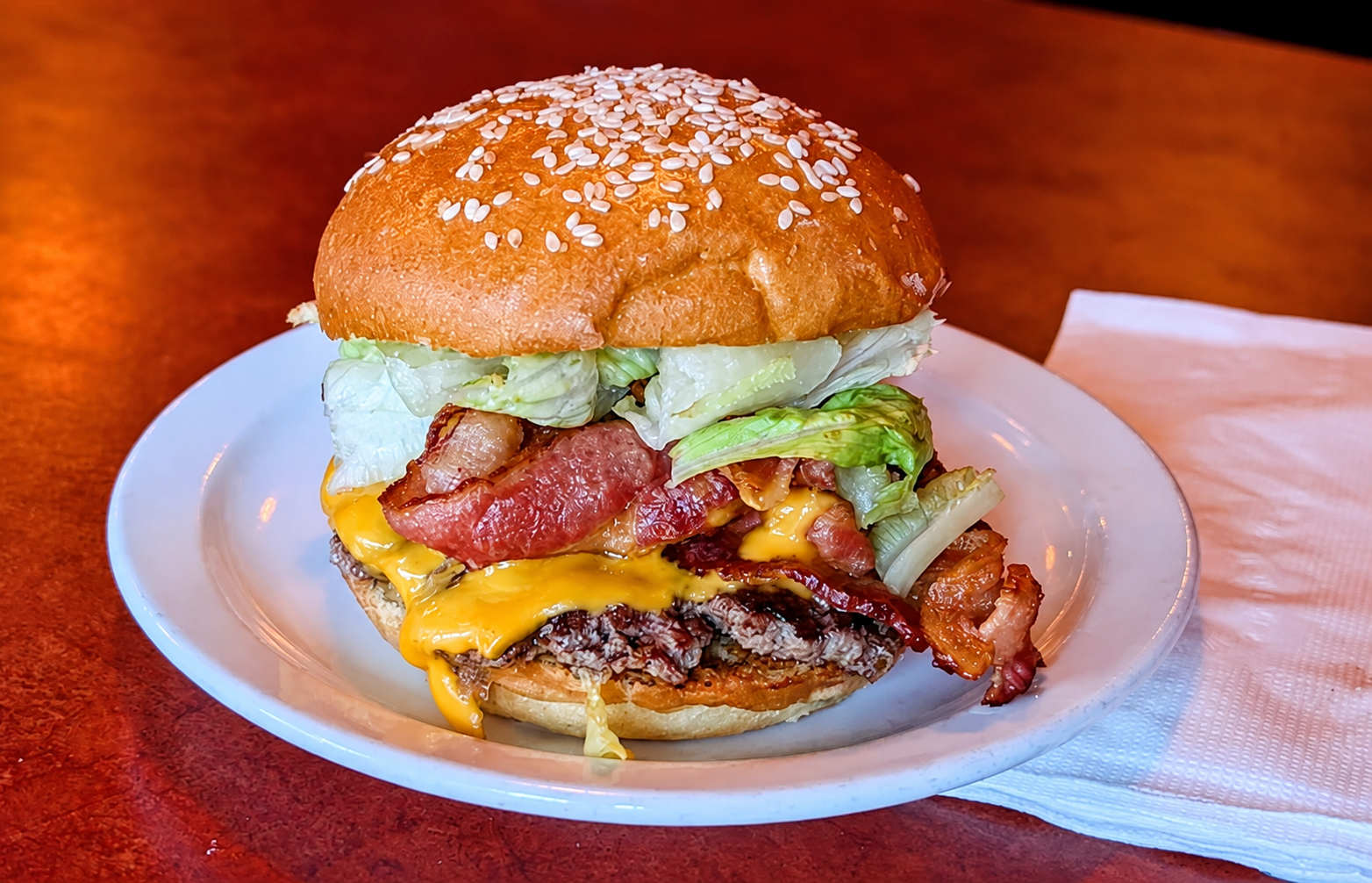 A cheeseburger with bacon, lettuce, and other toppings on a sesame seed bun, served on a white plate.