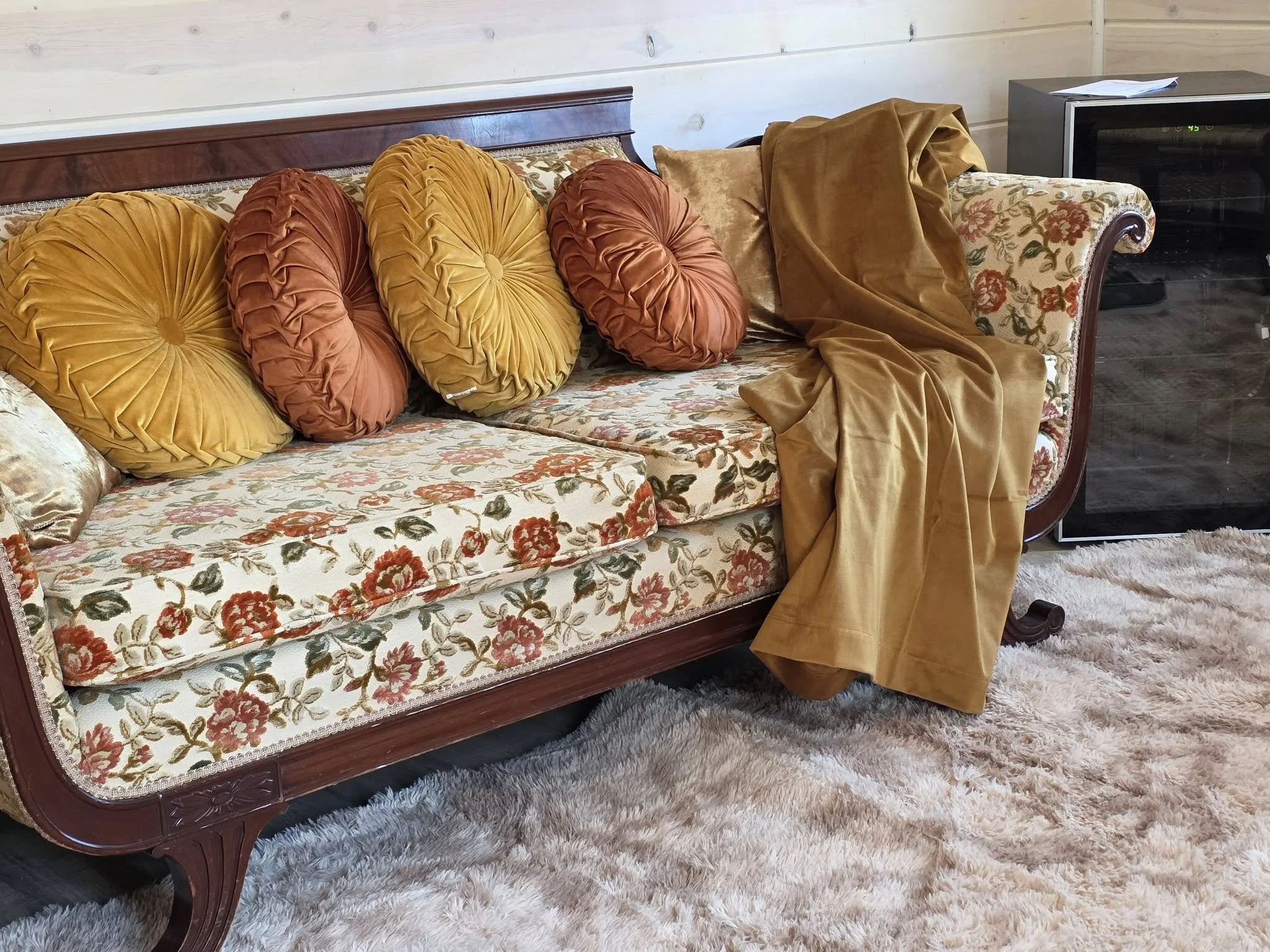 A vintage floral upholstered sofa with five large decorative pillows and a fur blanket, next to a mini refrigerator with champagne glasses on top, on a shaggy cream-colored rug.