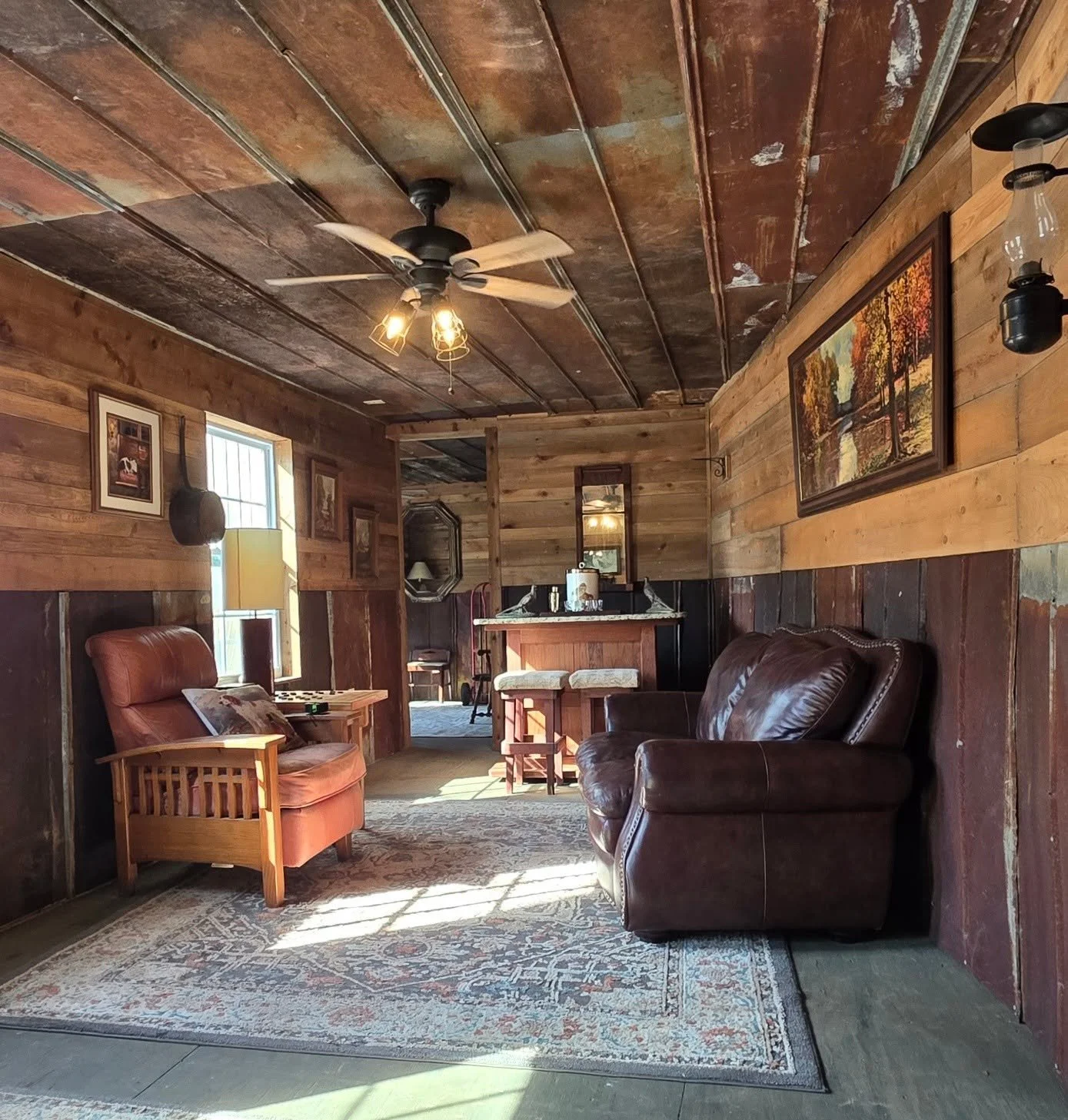 A cozy rustic living room with wooden walls and ceiling, featuring two leather armchairs, a patterned area rug, framed artwork on the walls, and a ceiling fan with lights.