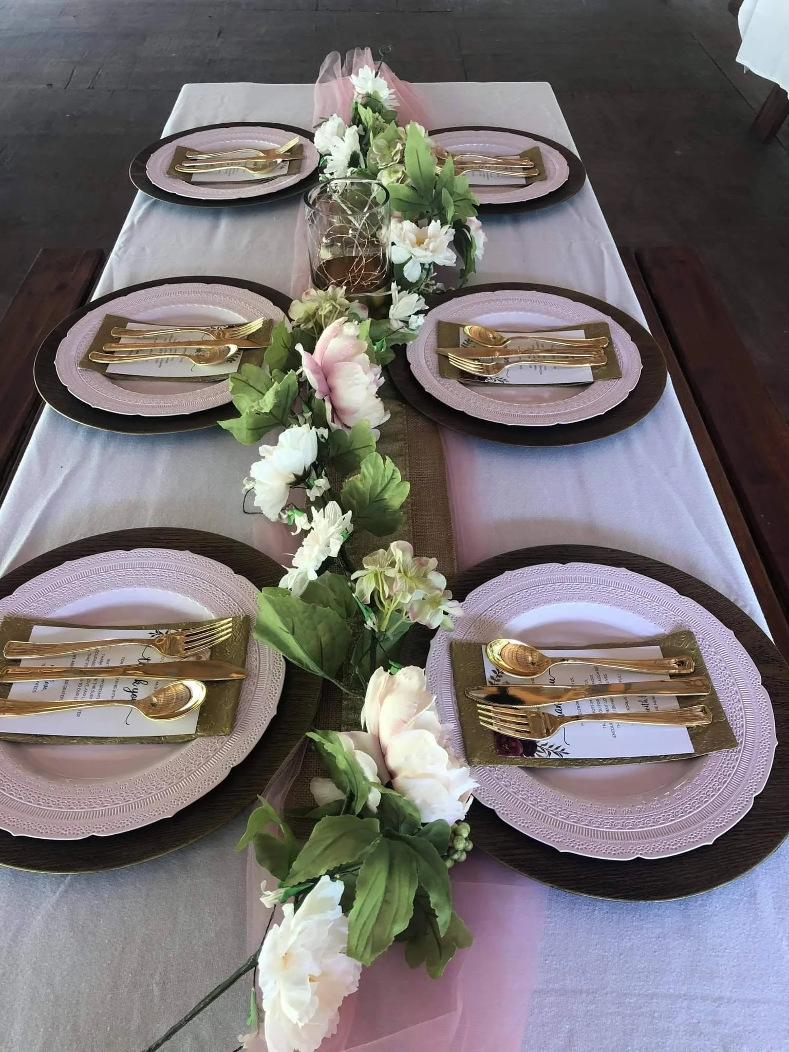 A dining table decorated with a pink table runner and floral centerpiece. Each setting includes a pink and brownplate, gold cutlery, and a menu card. The centerpiece features white flowers and green leaves, with some pink tulle fabric.