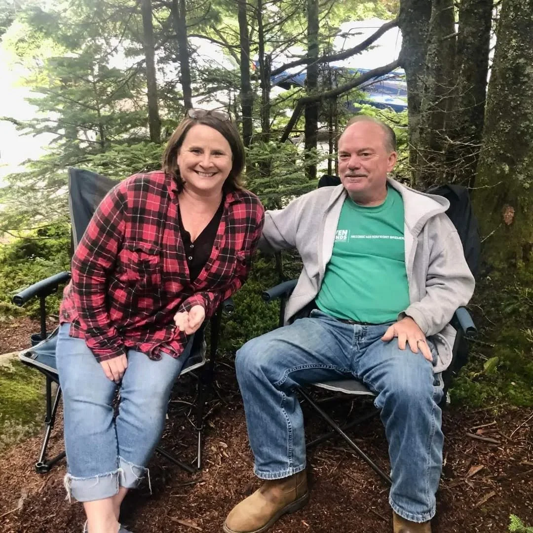 A smiling woman and a man sitting in camping chairs outdoors in a forest setting.