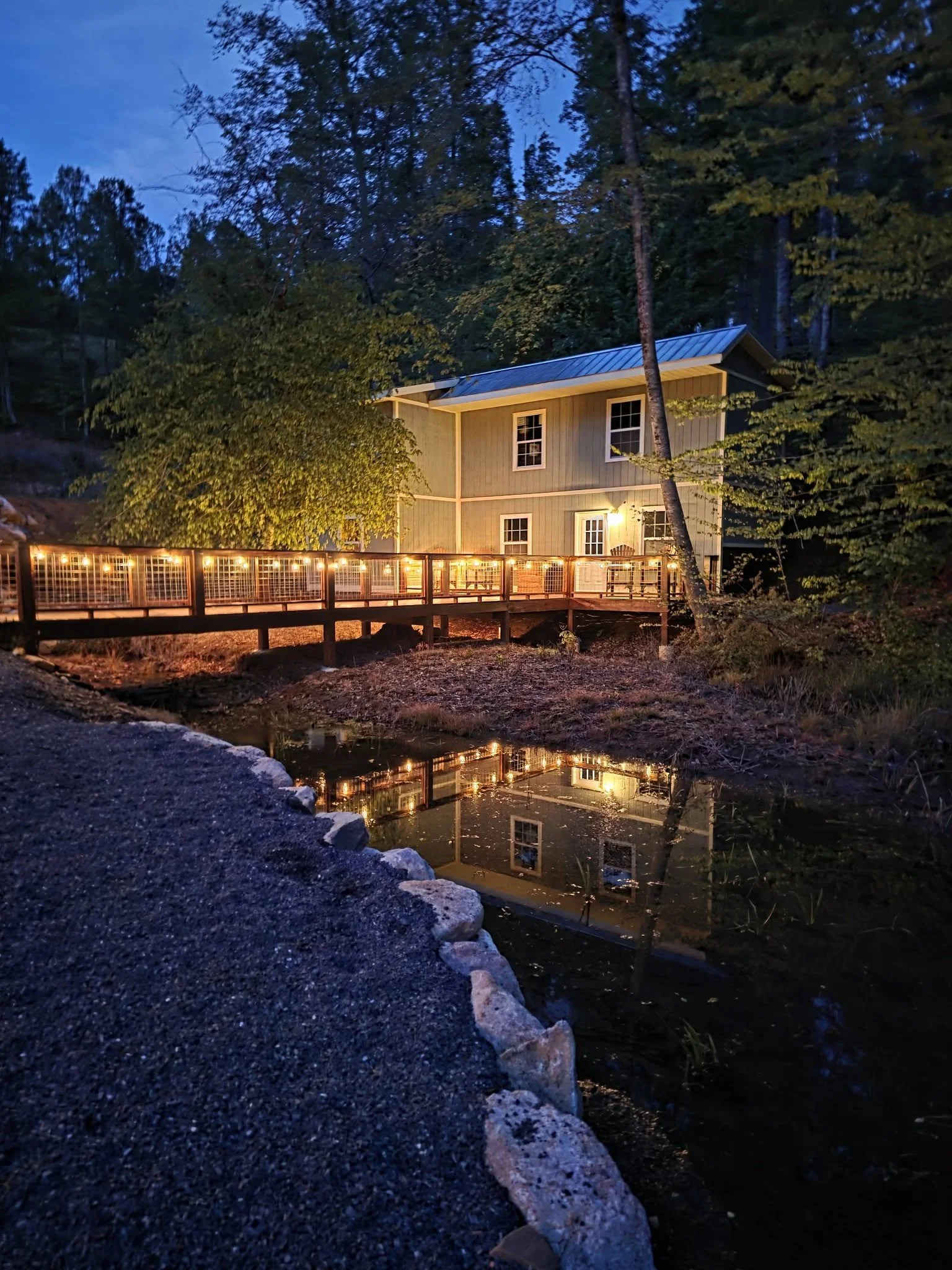 A cabin with a green exterior and white window frames situated by a small pond, illuminated by outdoor lights at dusk amid tall trees.