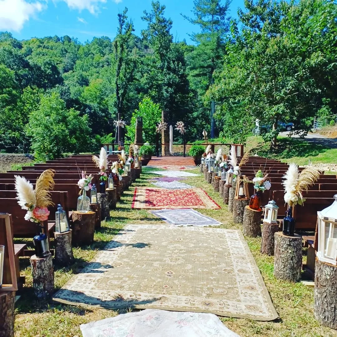 Outdoor wedding ceremony set up with chairs on either side of a decorated aisle, featuring floral arrangements, lanterns, and rugs, in a green, wooded area under a blue sky.