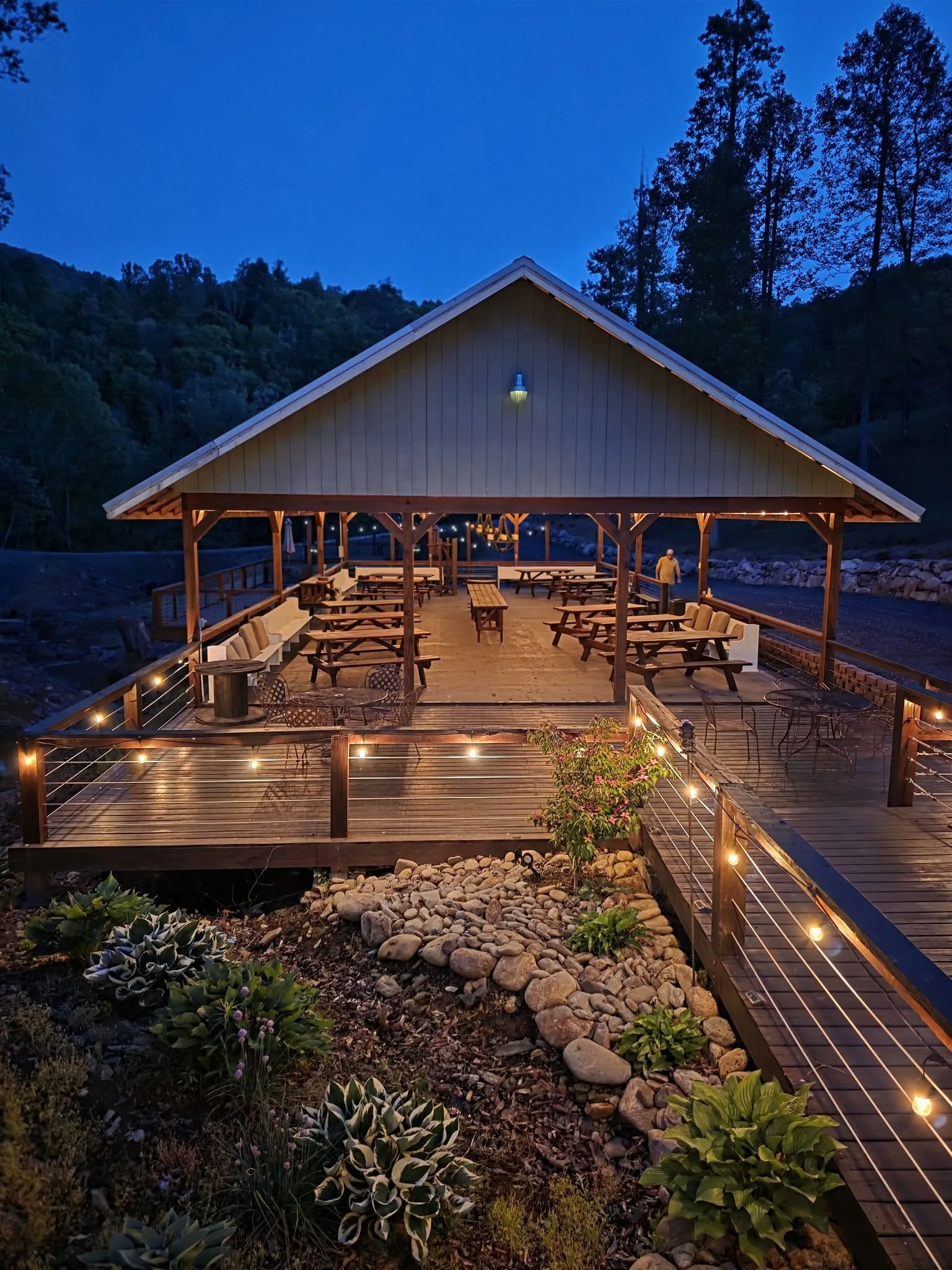 A covered outdoor pavillion with wooden railings and string lights, overlooking a rocky landscape with plants, on a dusky evening with the river in the background.