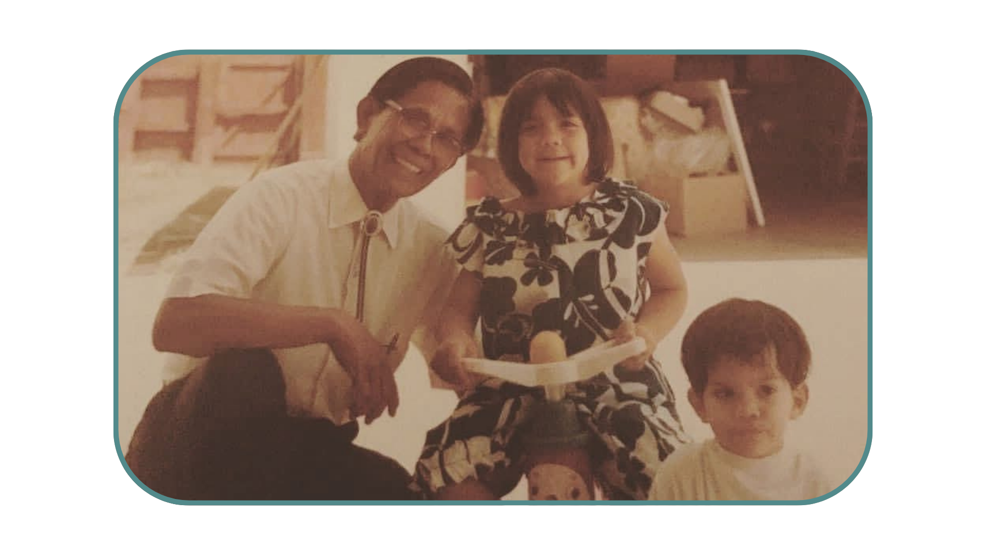 A man with short dark hair, glasses, and wearing a white shirt, smiling while holding a young girl with shoulder-length dark hair and a black and white patterned dress. A boy with short hair and a light-colored shirt is also partially visible in the foreground.