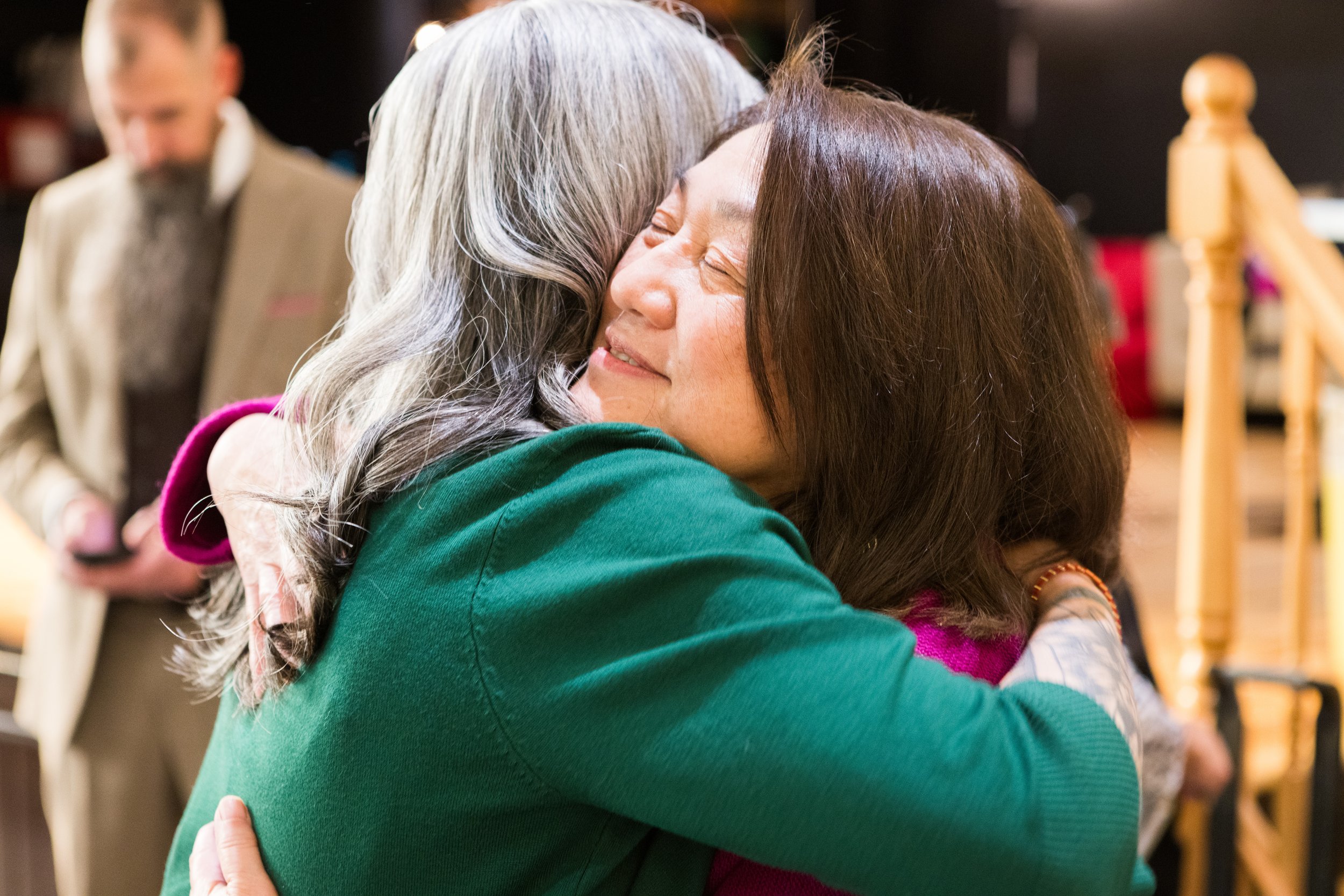 Two women are hugging each other tightly, smiling and showing affection. The background shows other people, possibly at a social gathering or event.