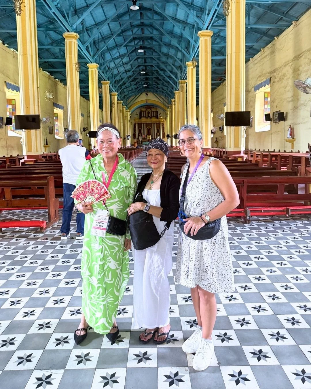 Three women standing inside a church with yellow walls and blue ceiling, wooden pews, and patterned tile floor. They are smiling at the camera, dressed in casual summer clothing, with one woman holding a pink fan. Other people are visible in the background.