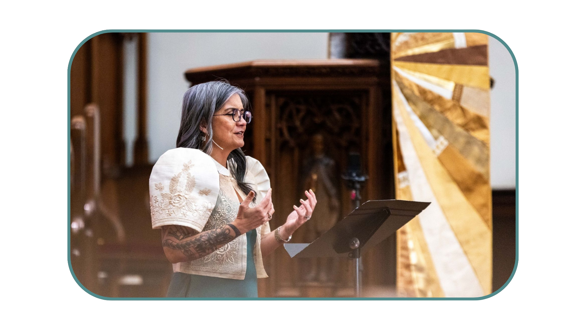 A woman with gray hair and glasses speaks at a podium in a church or hall with ornate woodwork and a colorful abstract wooden art installation behind her.