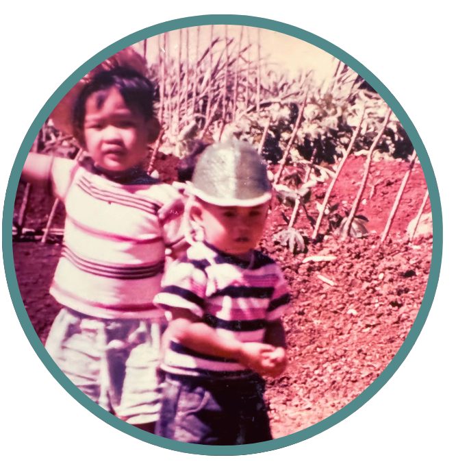 Two young children standing outdoors in a garden or farm, with one child wearing a striped shirt and a hat, and the other child wearing a striped shirt without a hat.