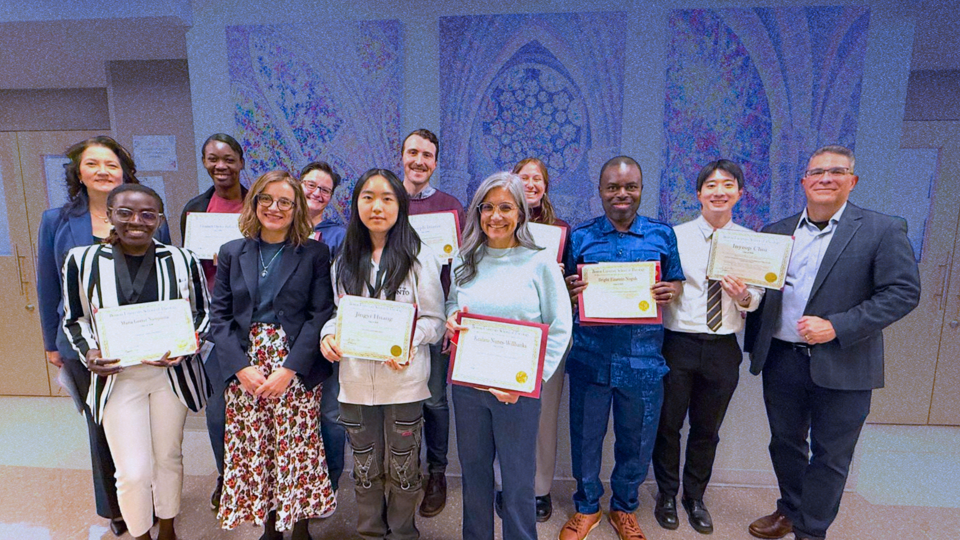 Group of diverse young adults and older adults holding certificates and smiling at an awards ceremony or graduation event in a decorated hall.