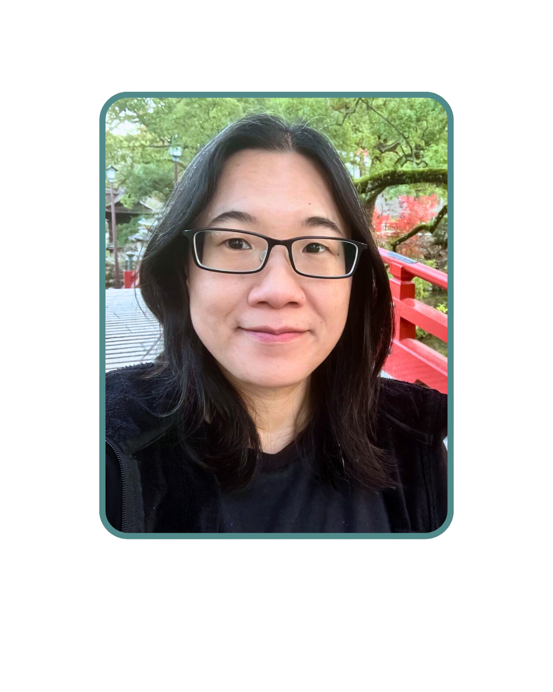 A woman with glasses and black hair taking a selfie outdoors on a bridge with greenery and trees in the background.