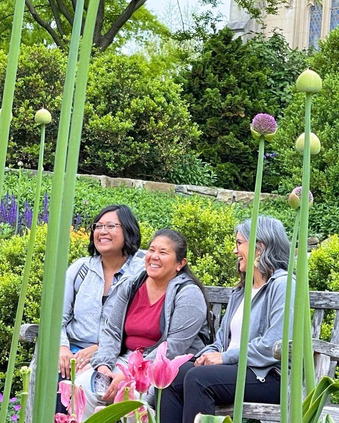Three women sitting on a wooden bench in a lush garden with blooming flowers and green foliage, enjoying a conversation and smiling.