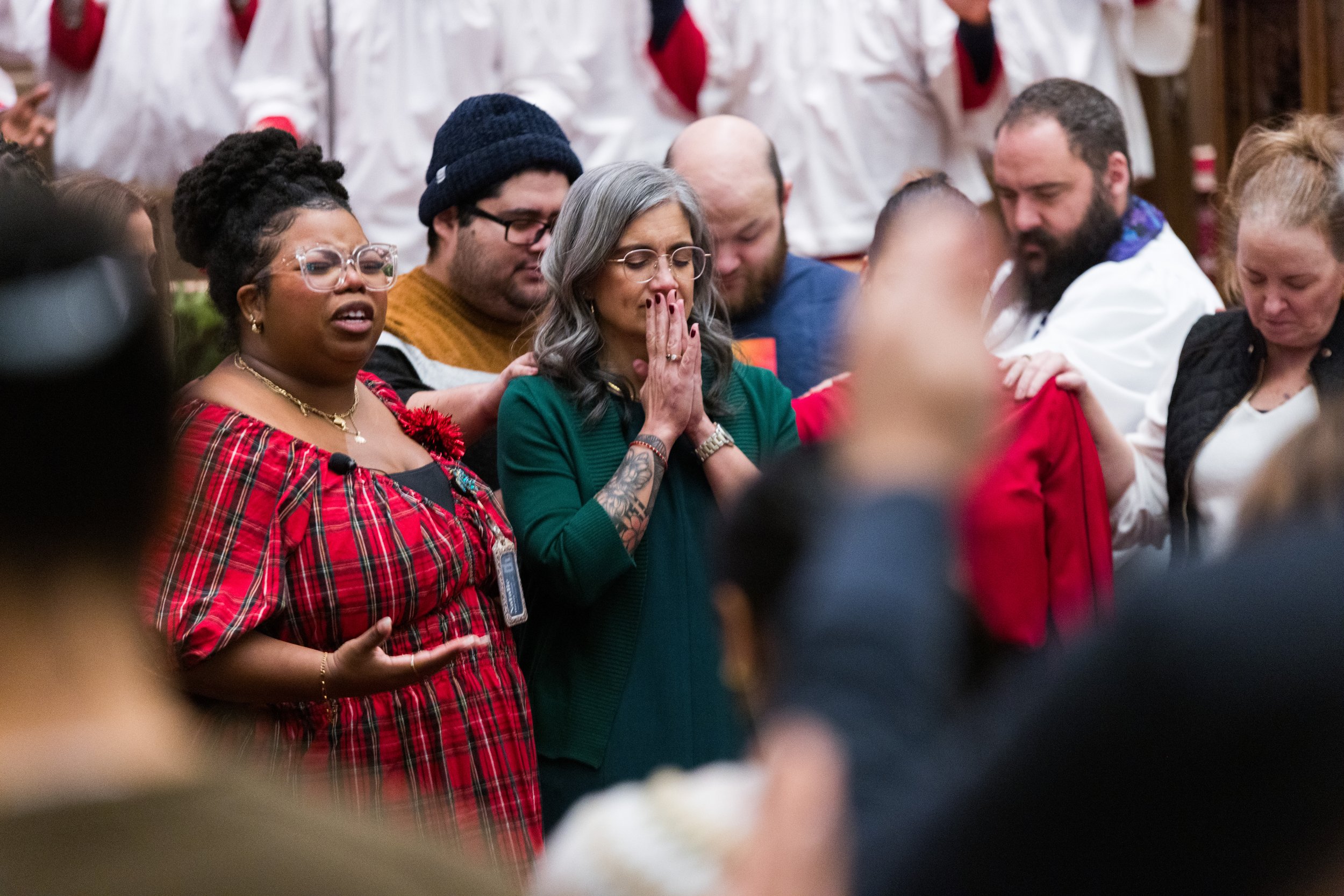 People participating in a prayer circle, with some holding hands and others with eyes closed, in an indoor setting.