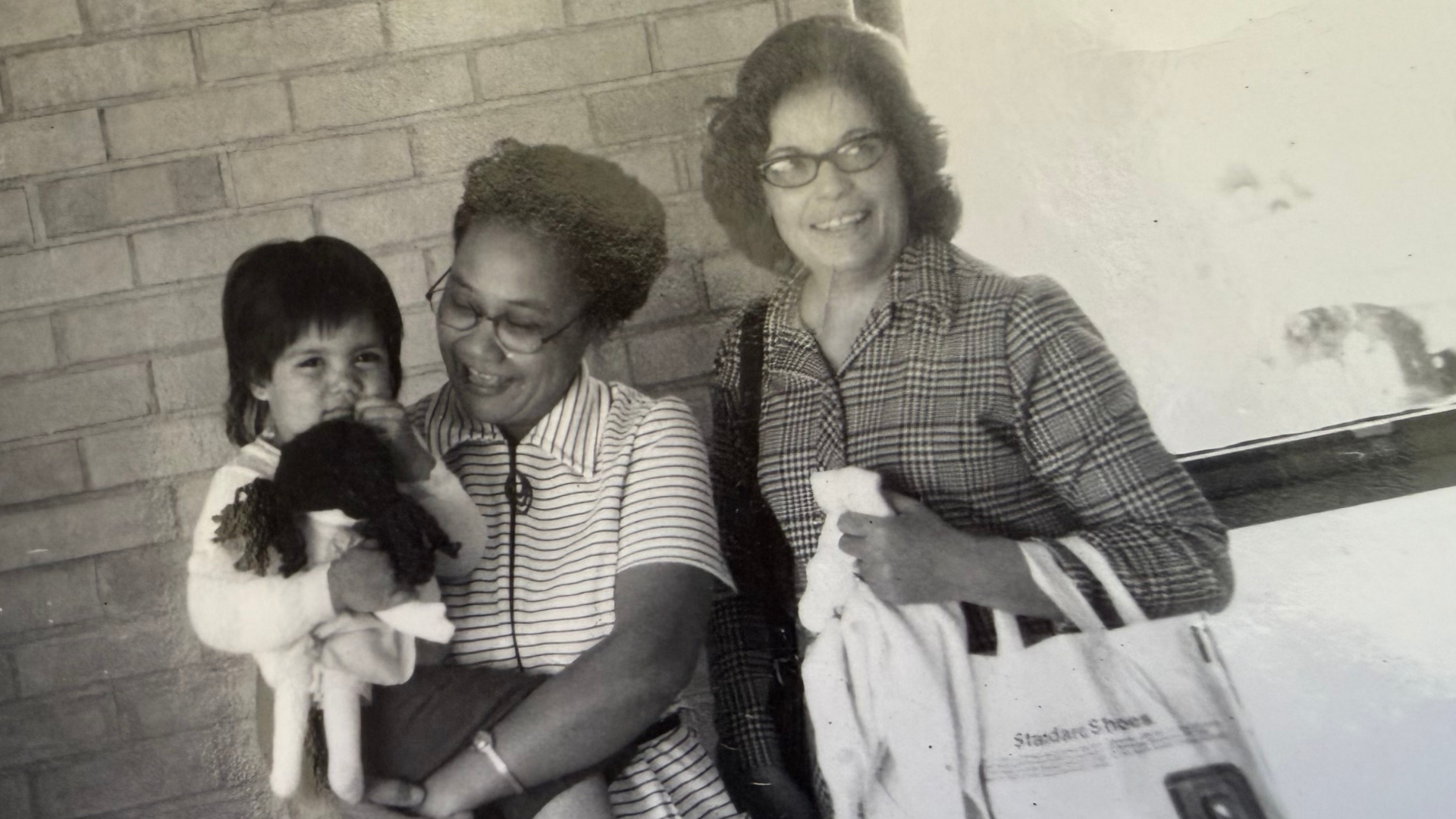 Three people, including a young girl and two women, sitting together against a brick wall. The girl has dark hair and is holding a stuffed animal. One woman, with glasses and short curly hair, is smiling and wearing a striped shirt. The other woman, with glasses and shoulder-length hair, is smiling and wearing a checkered shirt. They appear to be happy and enjoying each other's company.