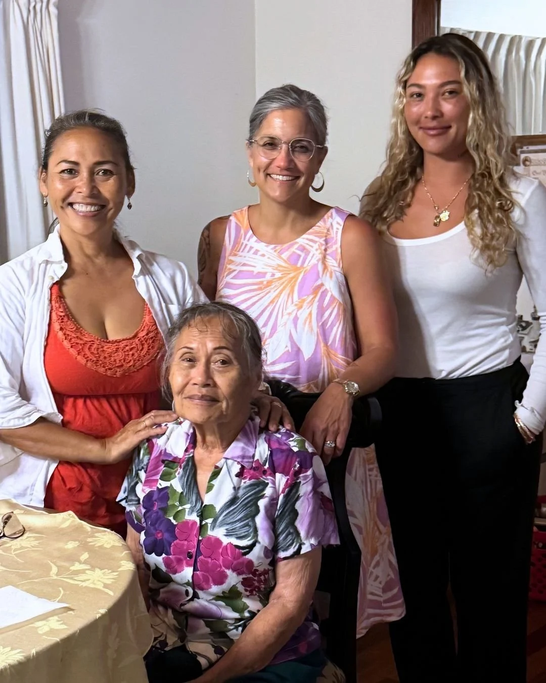 Four Filipina women, one elderly, gathered in a room smiling at the camera. One elderly woman is seated in front, wearing a floral shirt. The three other women stand behind her, dressed casually, with warm expressions and friendly poses.