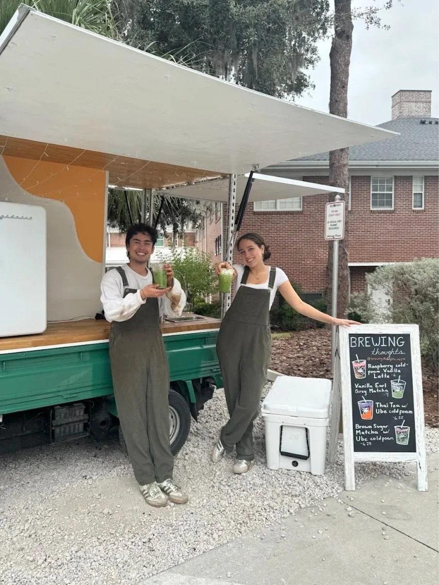 Two people smiling and holding green beverages in front of a mobile drink stand with a chalkboard menu listing various drinks, outdoors near residential buildings.