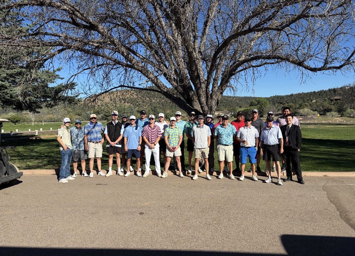 Group of people standing outdoors on a golf course in front of a large tree during daytime.