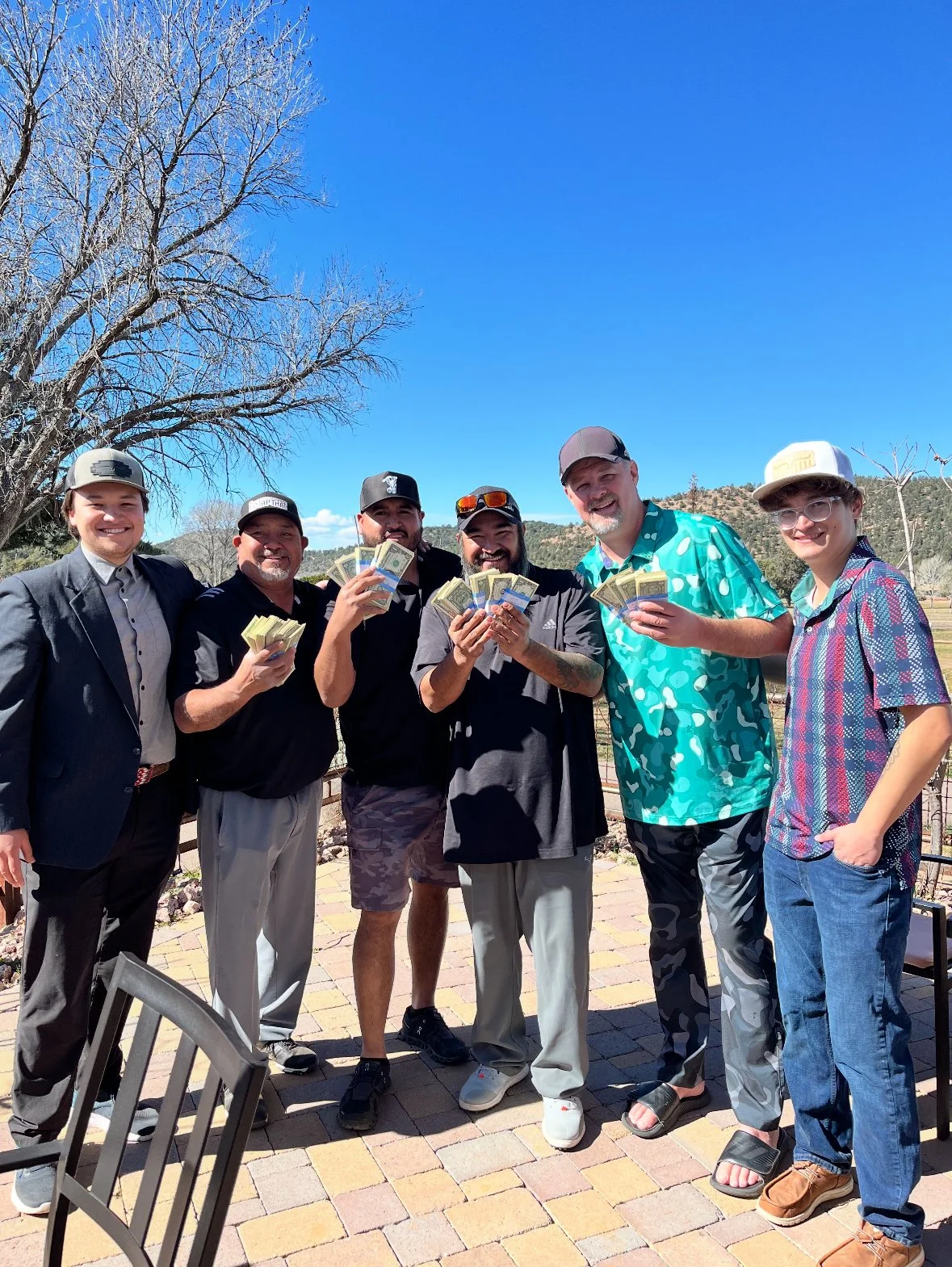Group of six men outdoors, smiling and holding stacks of cash, with a clear blue sky and leafless trees in the background.