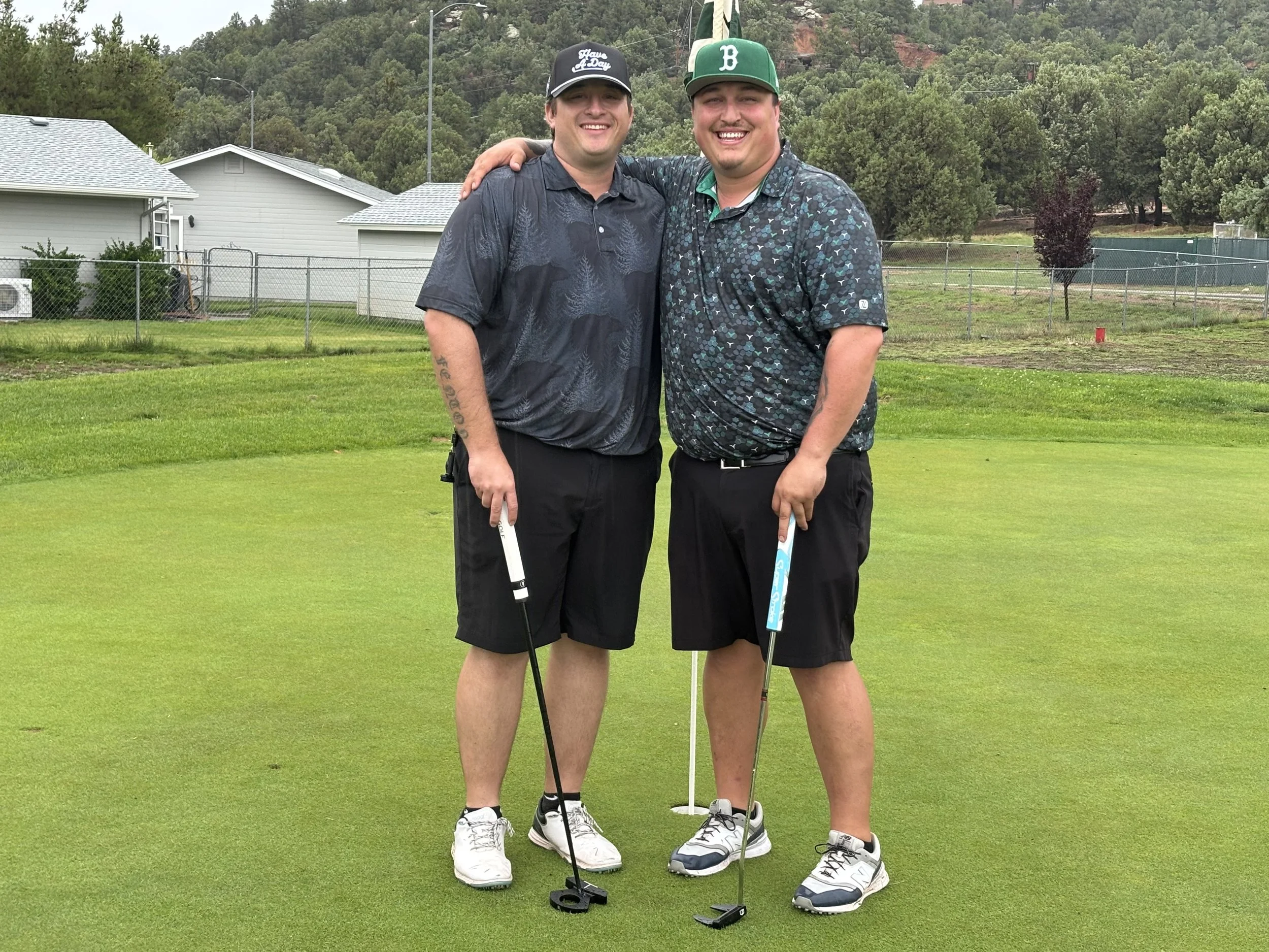 Two men standing on a golf course smiling, with one arm around the other's shoulder, holding golf clubs. They are dressed in golf attire, and there are houses and trees in the background.