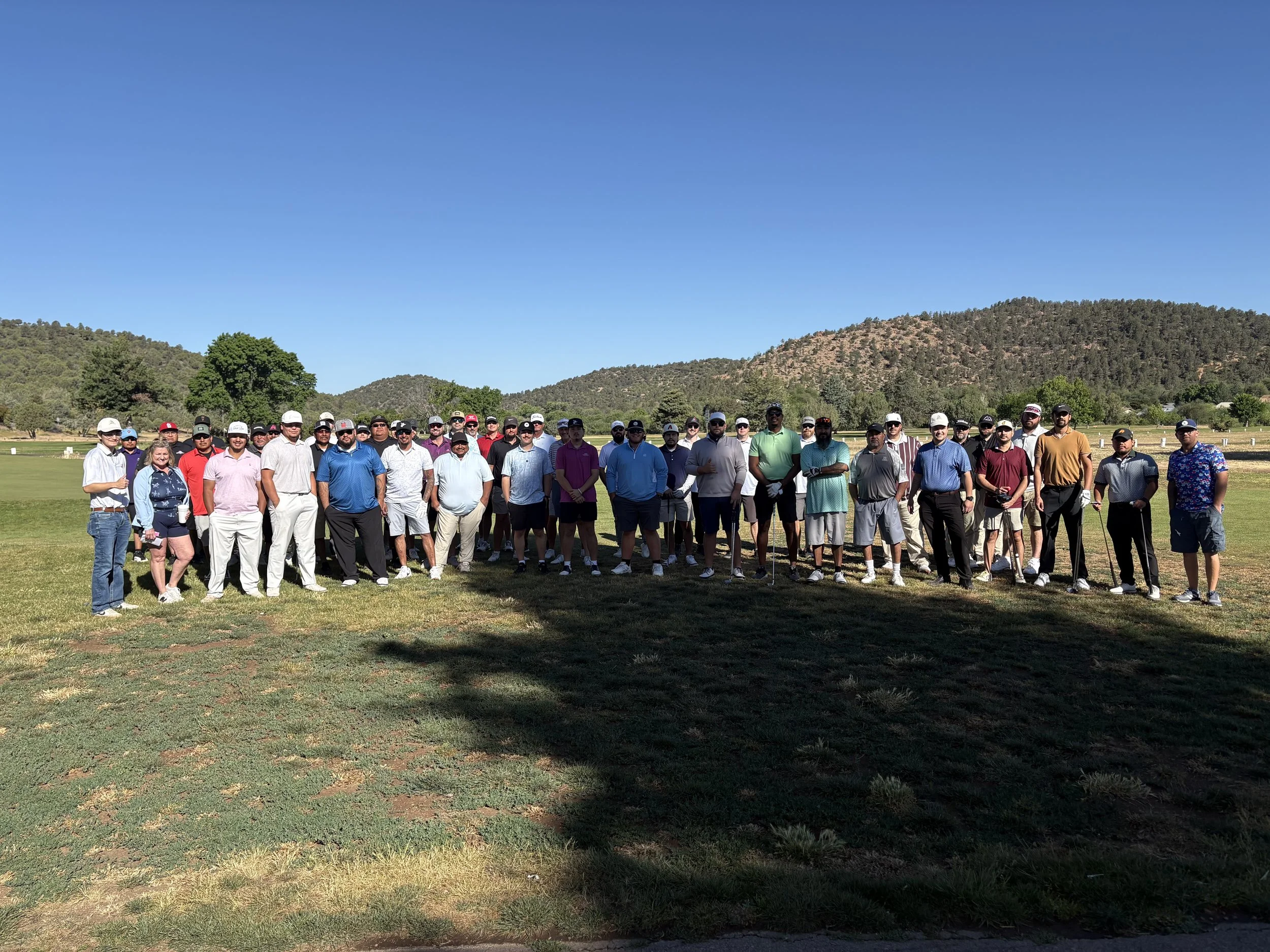 A large group of people standing outdoors on a golf course with hills and trees in the background. The sky is clear and sunny.