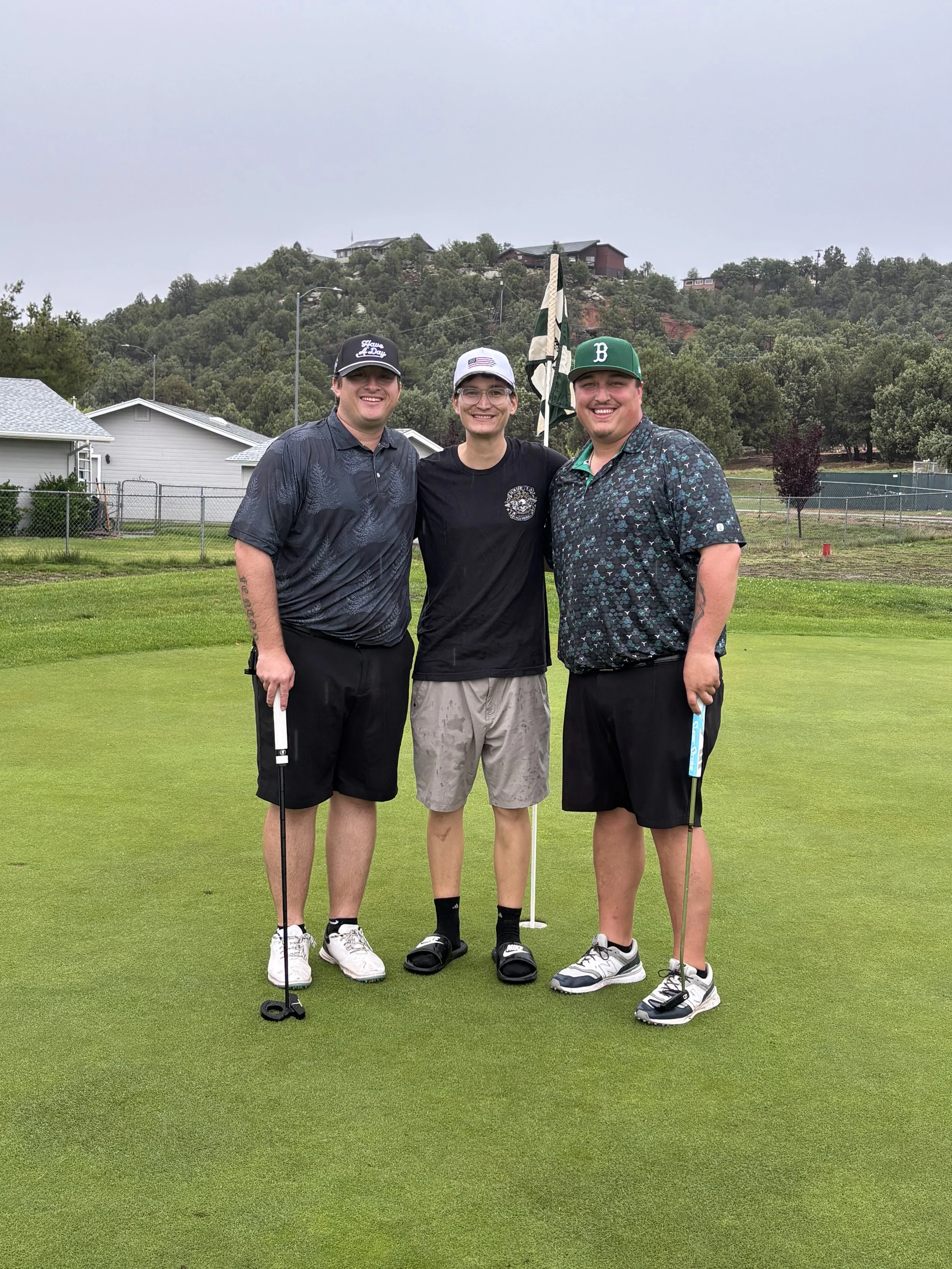 Three young men standing on a golf course green, smiling, holding golf clubs. Behind them are residential houses, a chain-link fence, and a hillside with trees and houses.